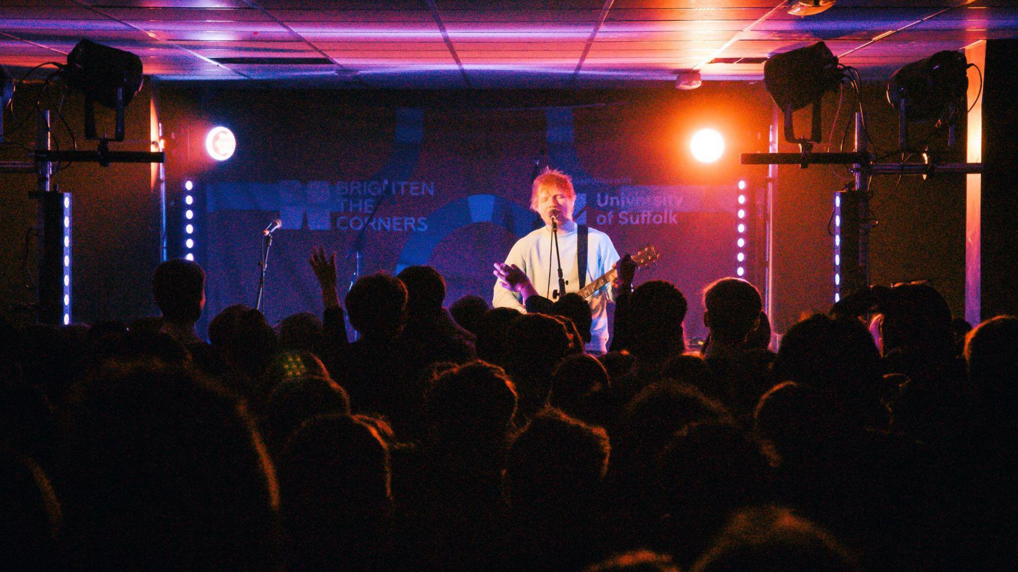 A concert sees man with guitar singing to crowd. It is dark and only silhouette of heads care seen. 