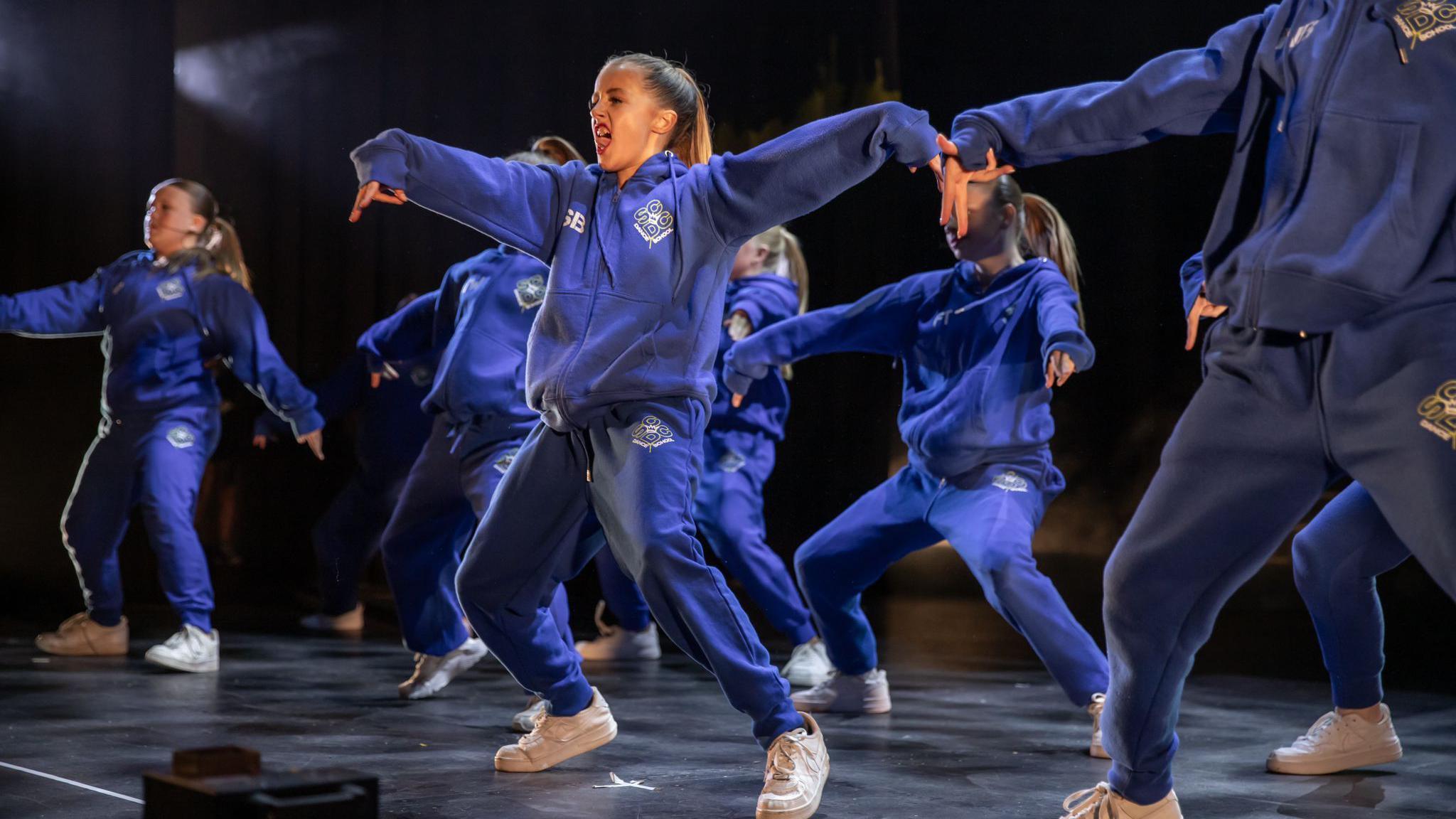 A group of children are dancing on a stage. They are wearing dark blue matching tracksuit bottoms and hoodies. Their arms are outstretched to the side.