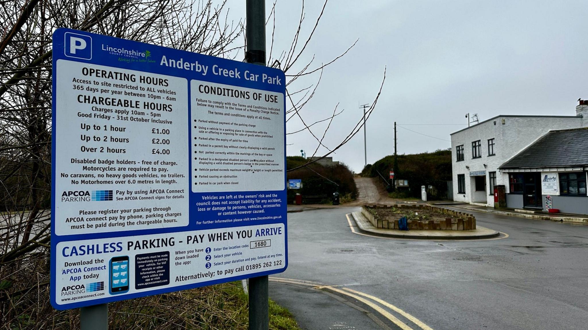 A large car park sign with methods of payment displayed. Behind is an empty car park with a white building in the corner.