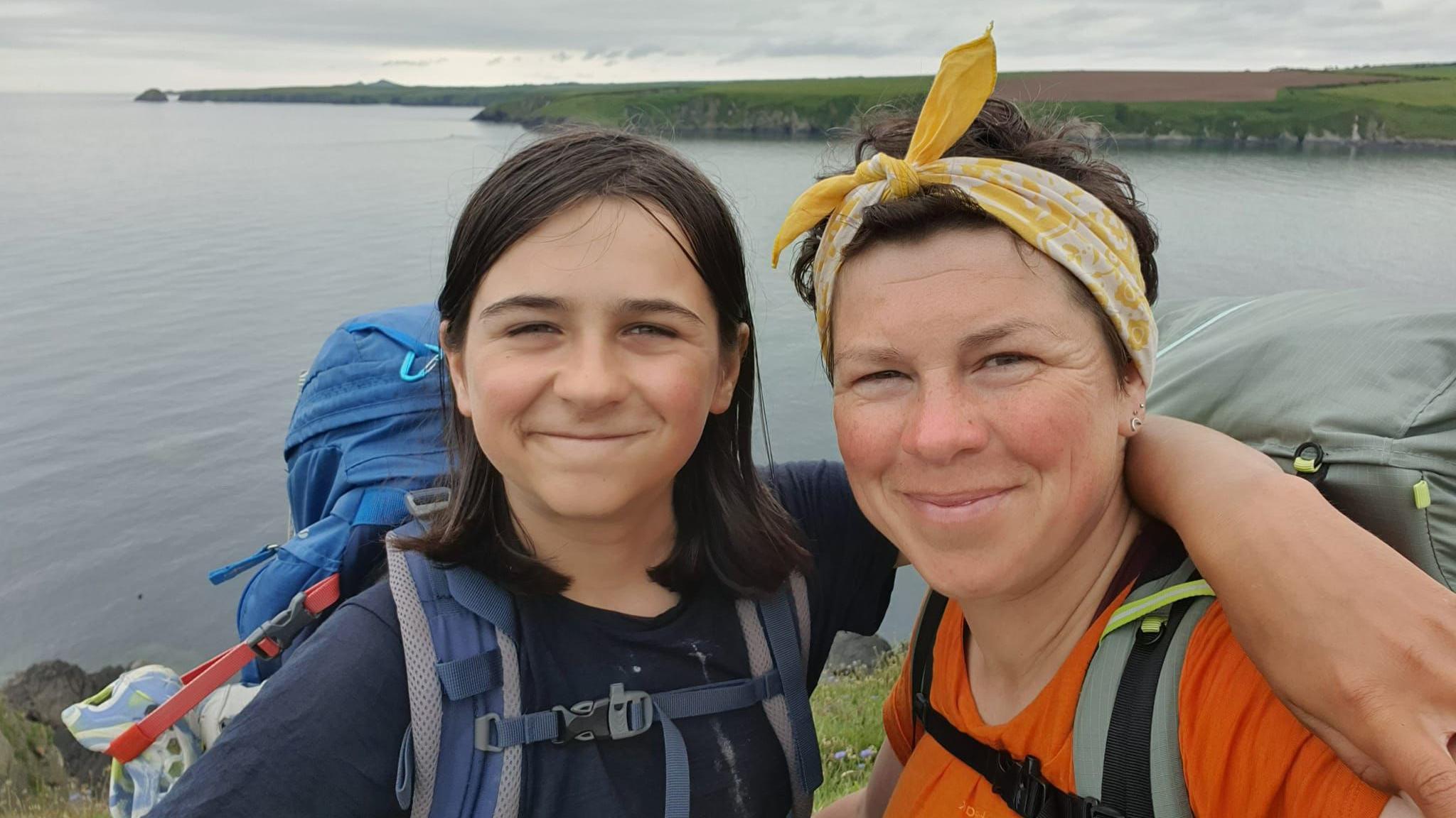 Finn and Kerry-Anne on the Wales Coast Path, they are both smiling and looking at the camera. Finn is wearing a blue t-shirt and Kerry-Anne is wearing an orange t-shirt and her hair is tied back in a yellow bandana