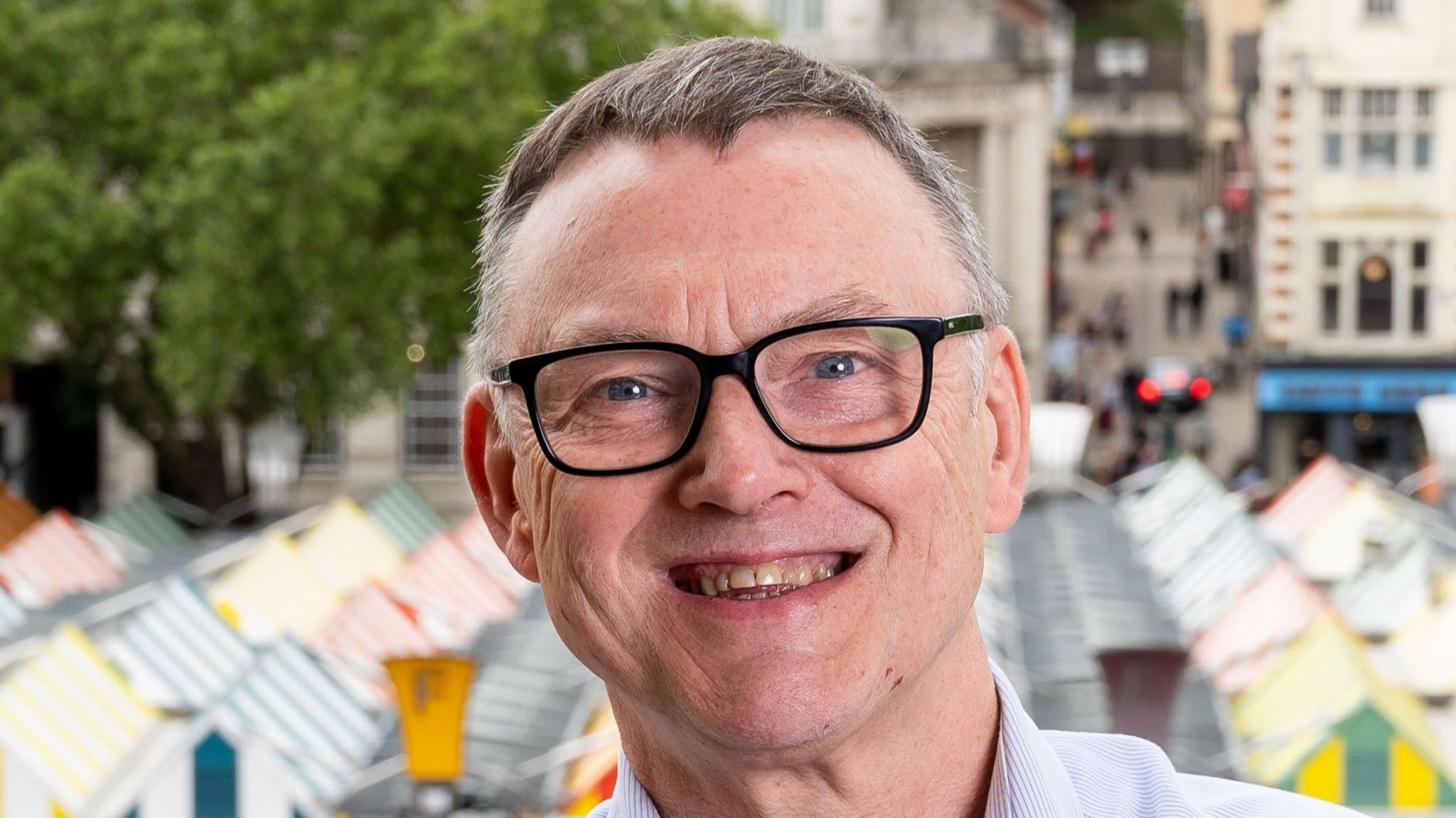 Mike Stonard, wearing a light-coloured shirt and black glasses, smiles at the camera. He has short grey hair. Behind and below him are the brightly-coloured canopies of Norwich Market's stalls. 