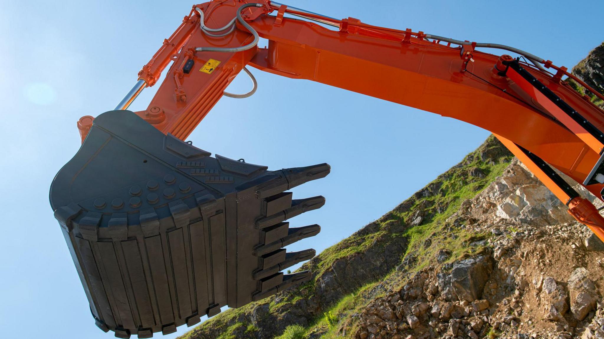 Close-up shot of a large orange excavator arm with a heavy-duty bucket. The excavator is on a hillside with blue sky in the background.