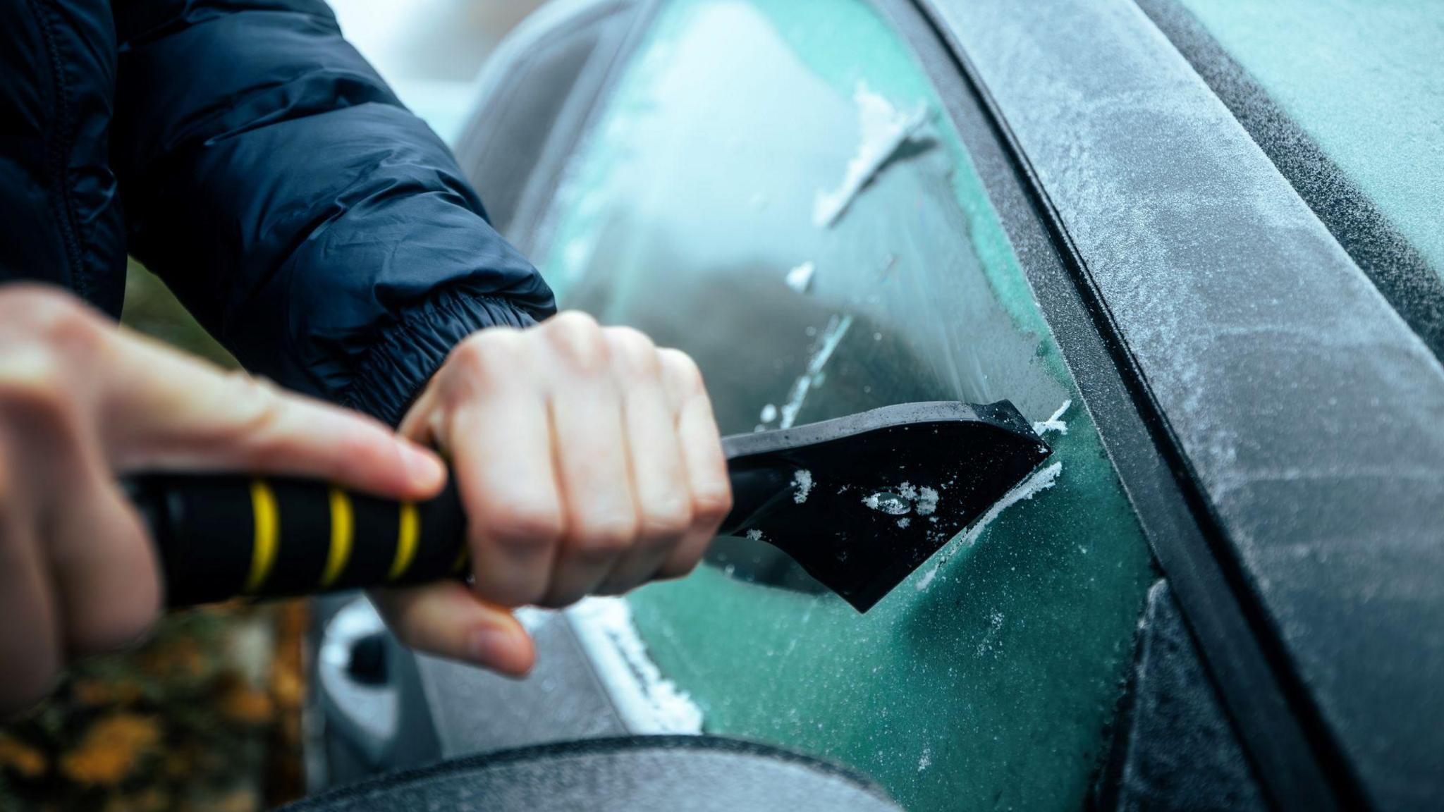 A person holds a black and yellow ice scraper as they work to remove ice from a car window.