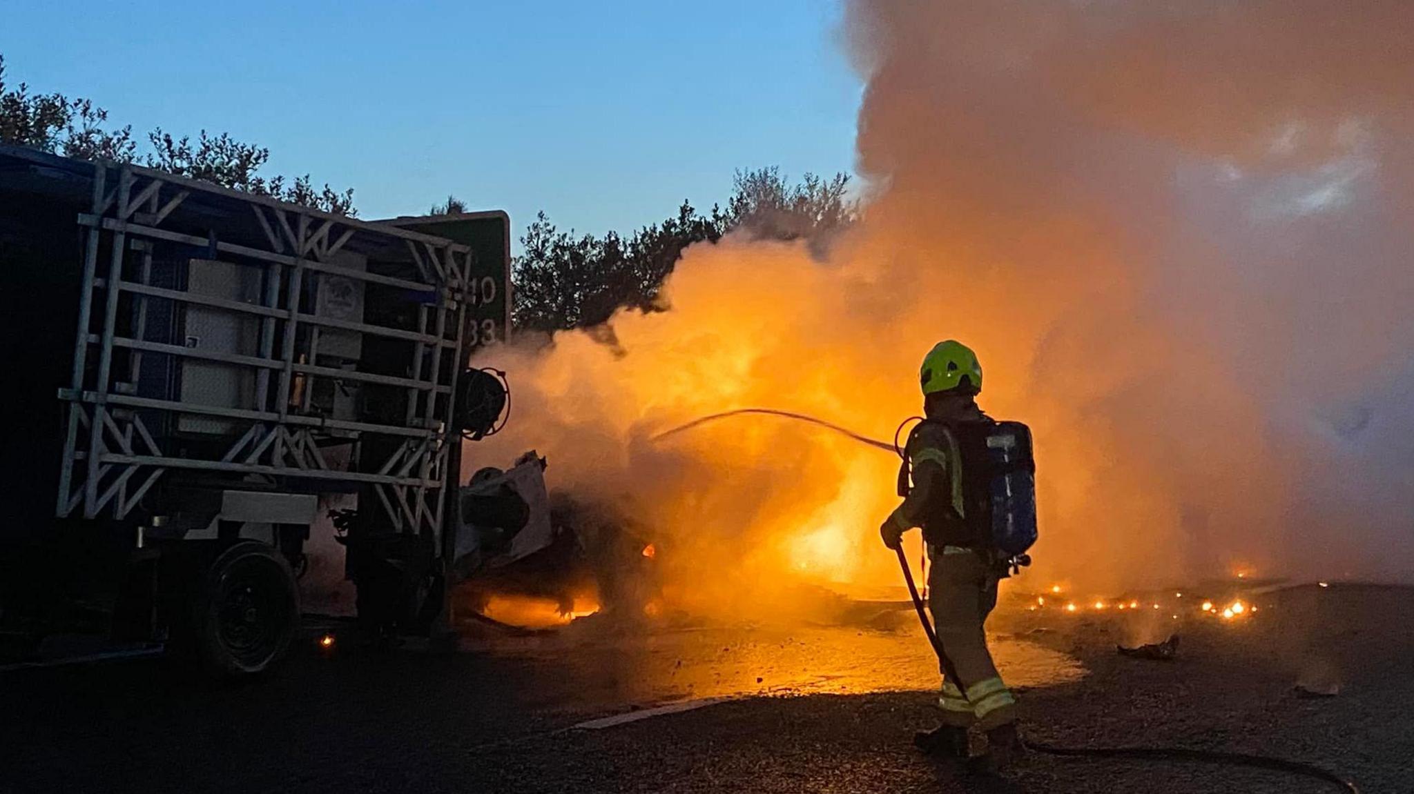 A firefighter spraying water on a vehicle on fire. There are large flames and smoke