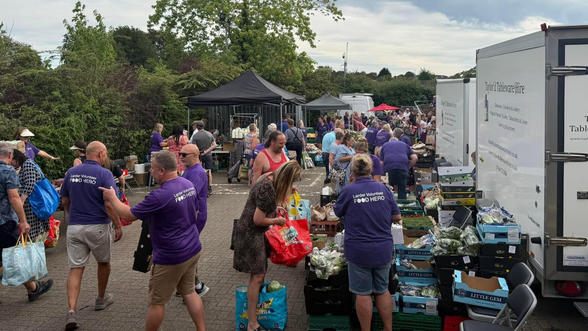 A large group of people at a community food larder, with gazebos on the left, vans to the right, and boxes of food at the front. Helpers are wearing purple T-shirts. There are trees in the distance. 