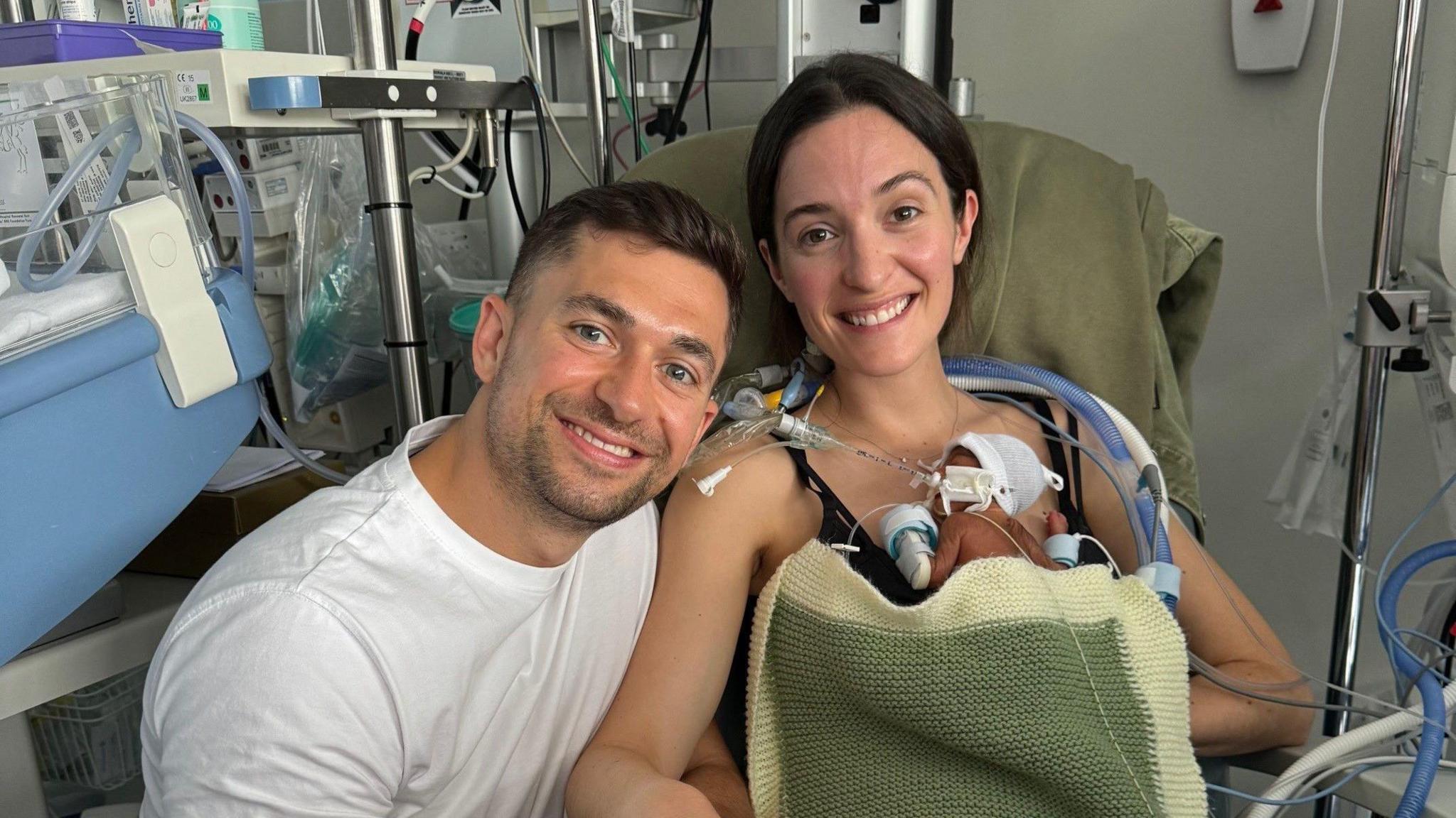 A dark haired young man sits in hospital with a dark haired young woman. The woman hold a small baby on her chest. The baby is wired to medical equipment.