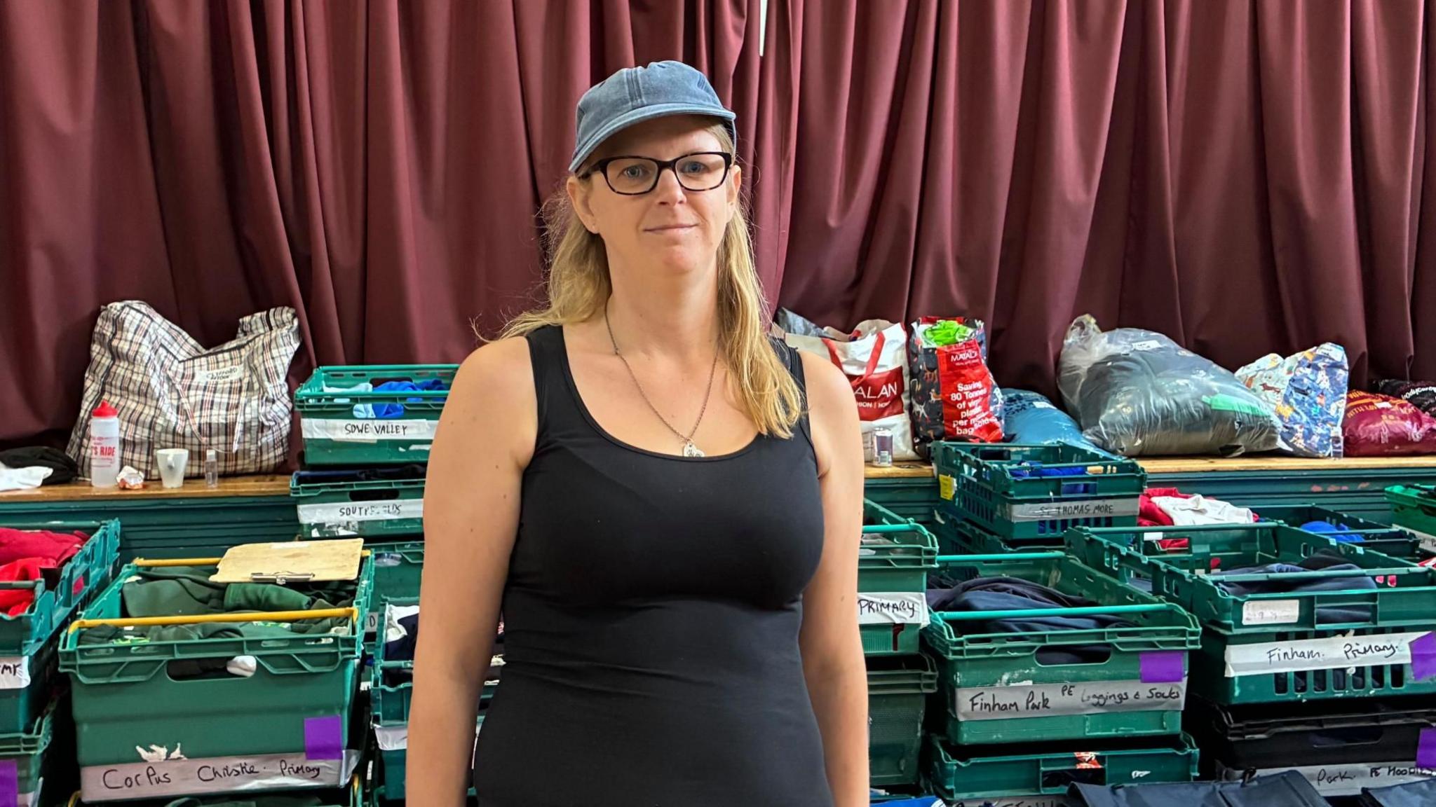 Zia Lee stands in a black vest top with a denim cap on and black glasses and long blonde hair. Behind her are green crates with school names and carefully folded uniforms in them.