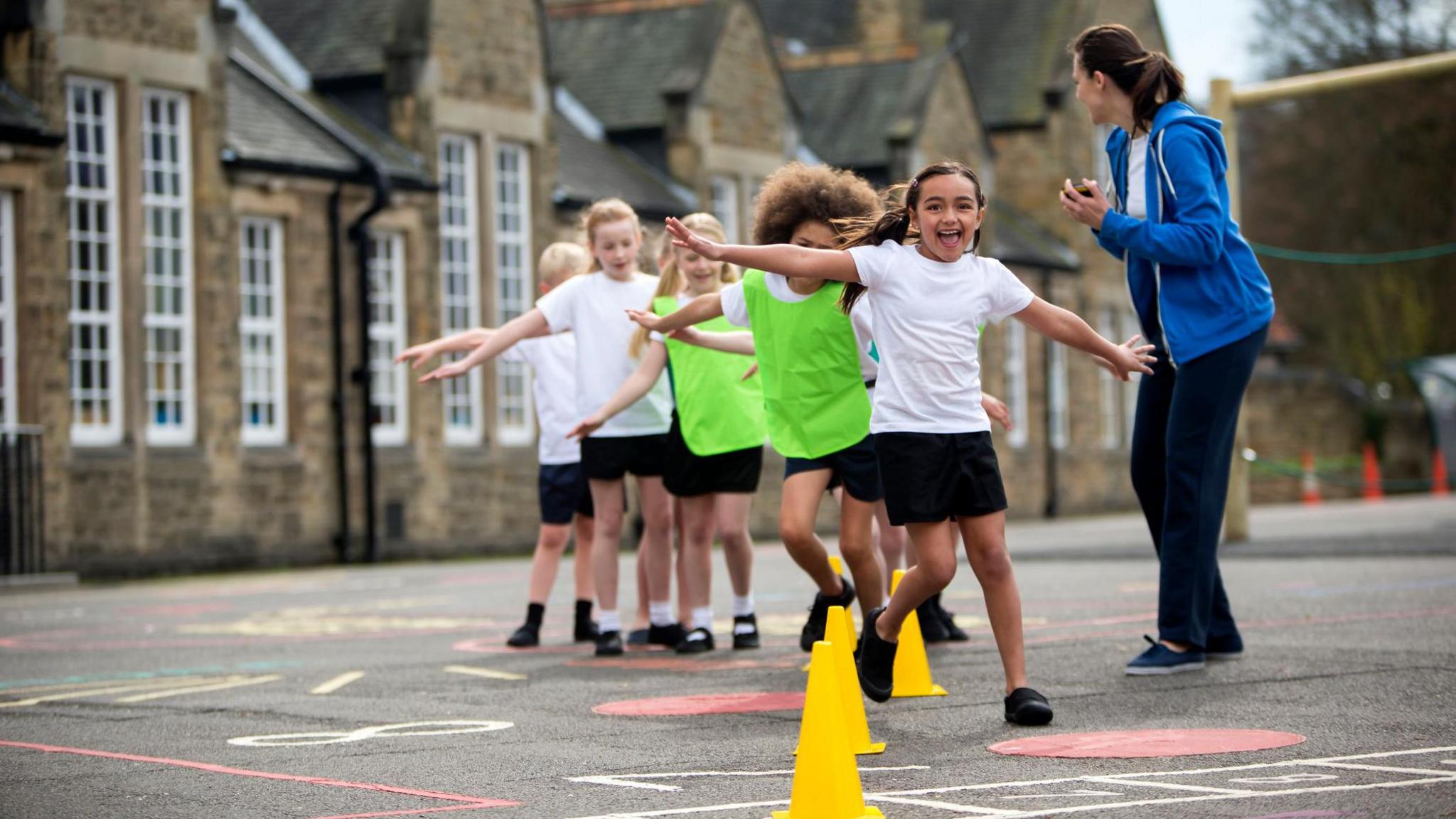 Children dressed in school PE kit exercising by running in and out of cones in a school playground, there is a teacher dressed in a blue hoodie next to them. 