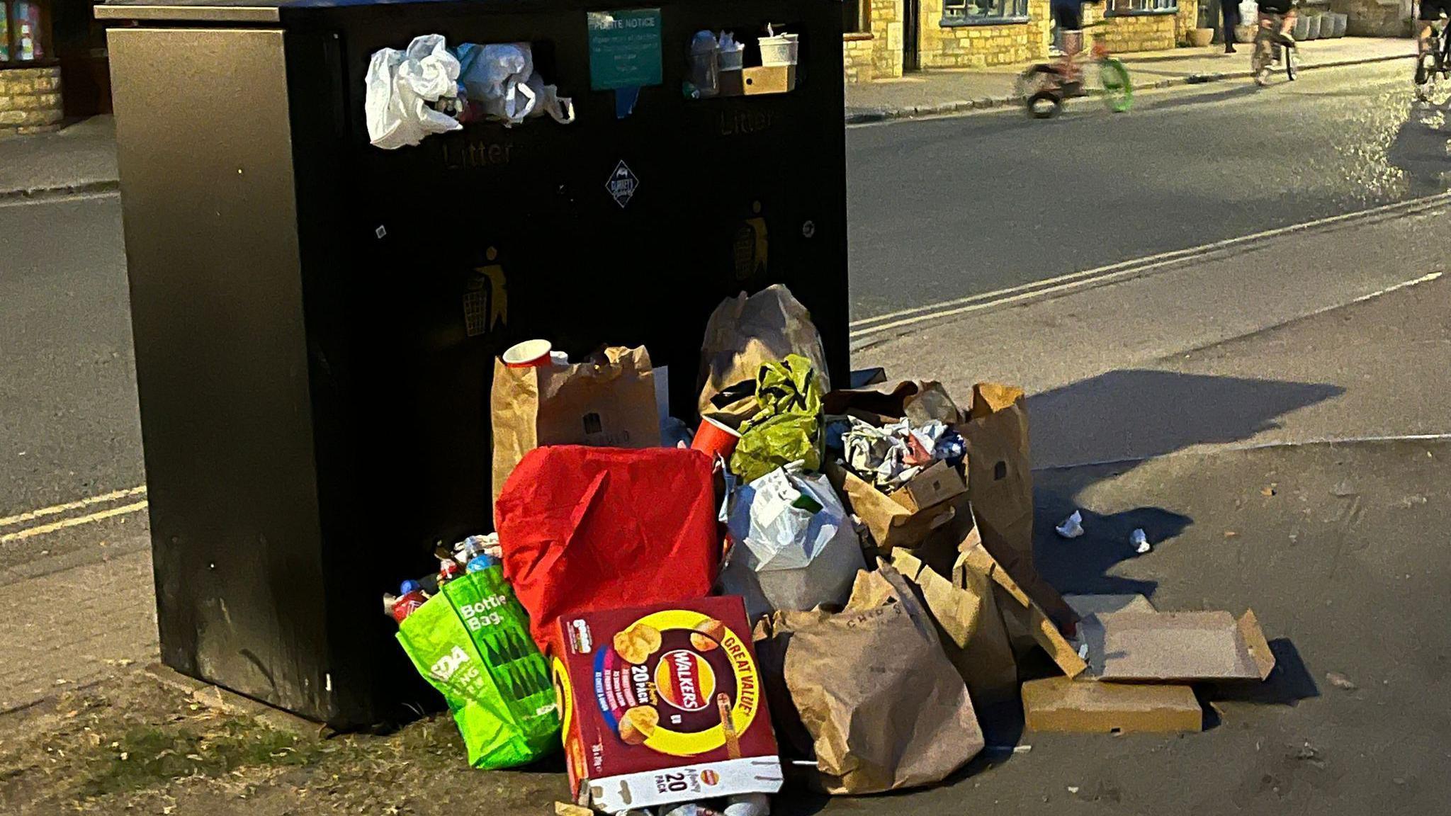 A pile of rubbish, including takeaway boxes and brown paper bags, and a box for crisps, piled next to a rubbish bin which is full up.