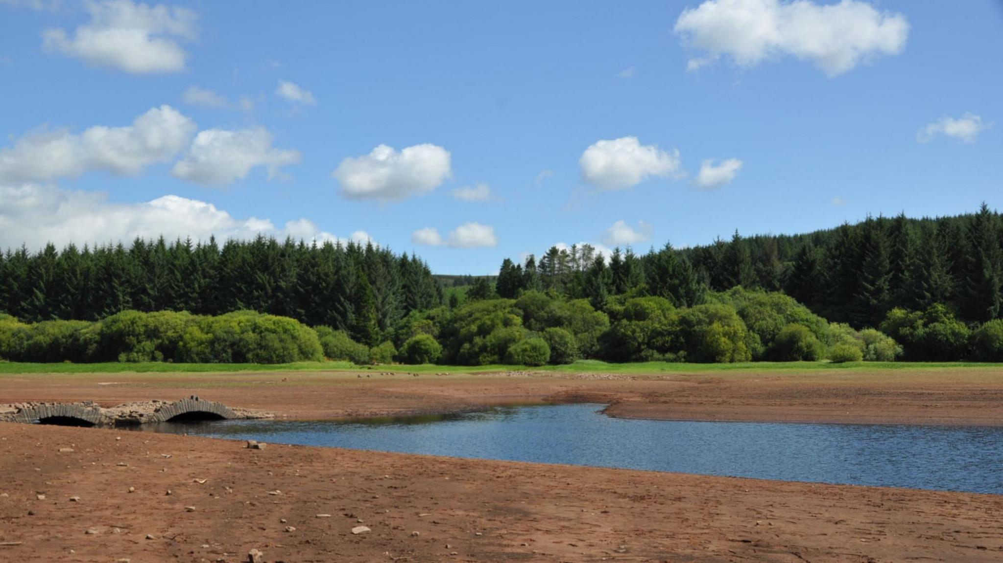 A dried out reservoir bed with a small amount of blue water and an old stone bridge. Green grass, trees and blue sky with clouds are in the background.