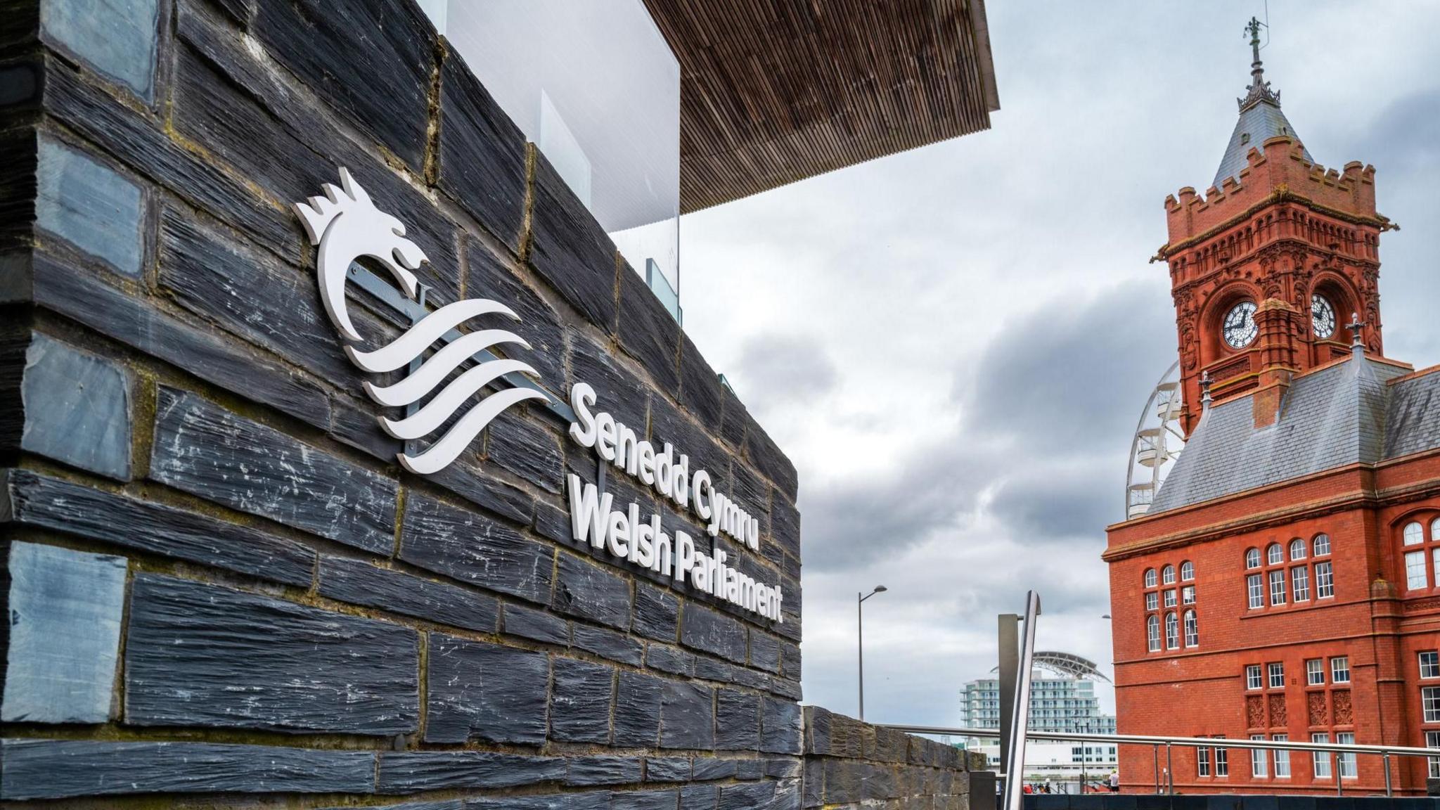 Llun o ran o adeilad y Senedd gyda enw'r a logo'r sefydliad. Ar ochr arall y llun mae modd gweld adeilad y Pierhead ym Mae Caerdydd.