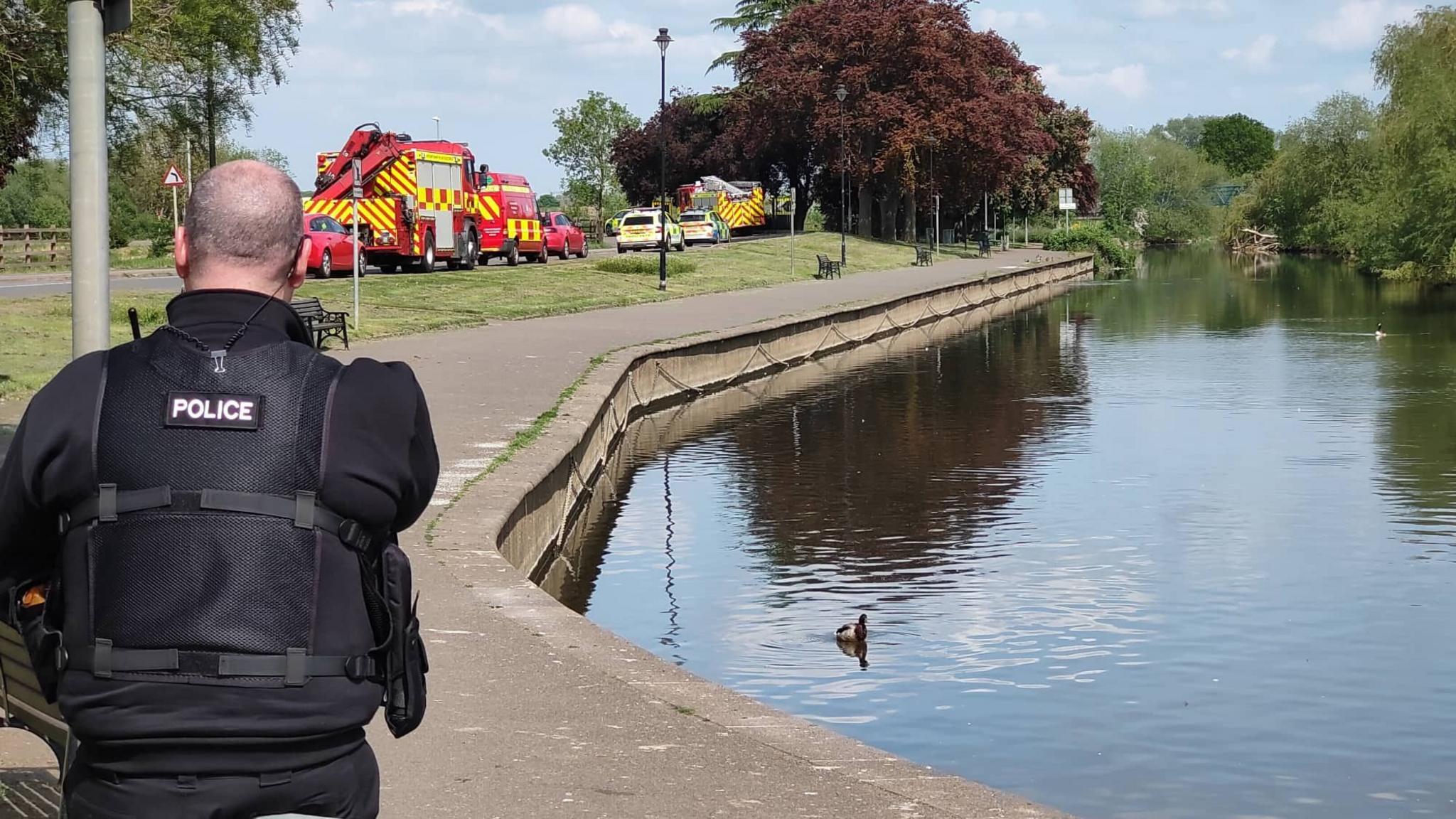 A police officer guards the River Nene at the embankment in Wellingborough. Rescue vehicles can be seen in the background.