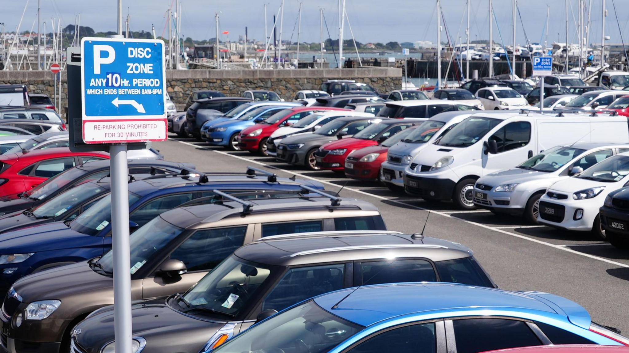Dozens of cars parked in car park in Guernsey. A blue sign saying the area is a "disc zone" is in the foreground. A marina with dozens of boats in the water is in the background.