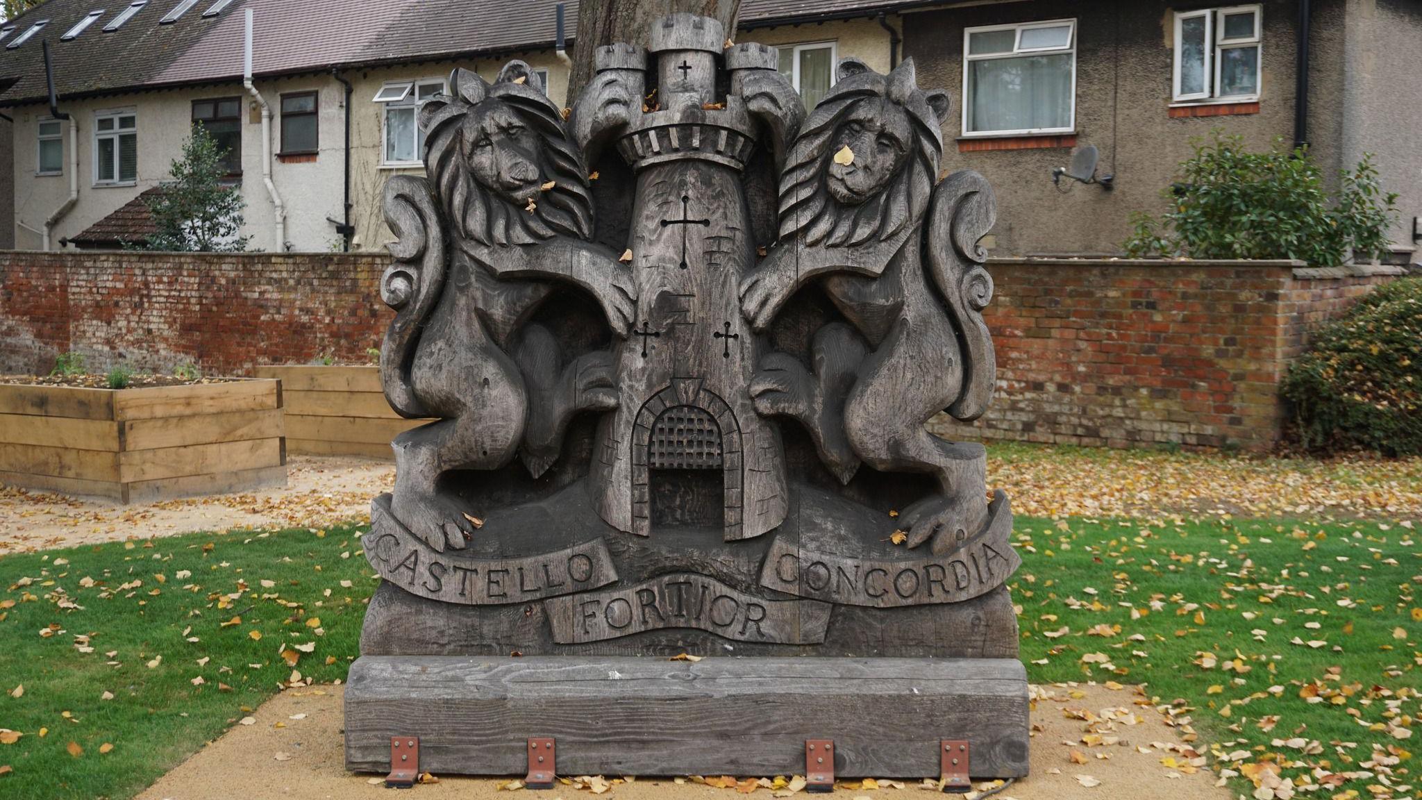 A large wooden coat of arms, in a park, showing two lions by a castle. There are leaves on the ground, a grass area and houses behind it.