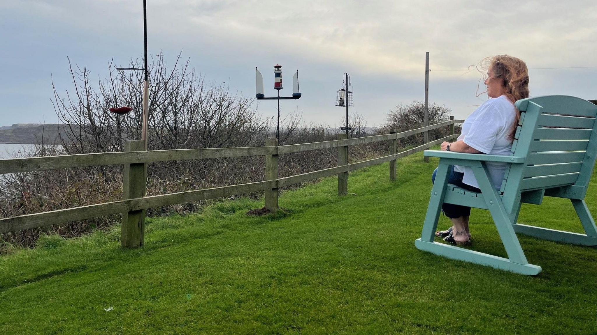 Angie Moore sits in a pale green, wooden chair on grass overlooking the sea. On the far left of the picture, is a headland.