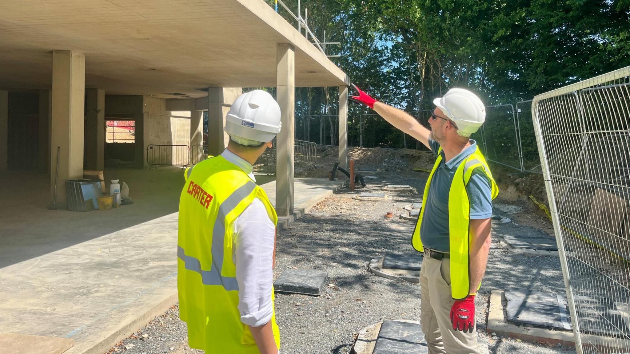 Two builders from RG Carter stand in front of the new extension to the Edith Cavell Building at the University of East Anglia in Norwich. They are wearing yellow tabbards and are pointing at the upper level of the building. In front of them you can see a concrete framework where the teaching suite will be. There is a metal safety fence to their left.
