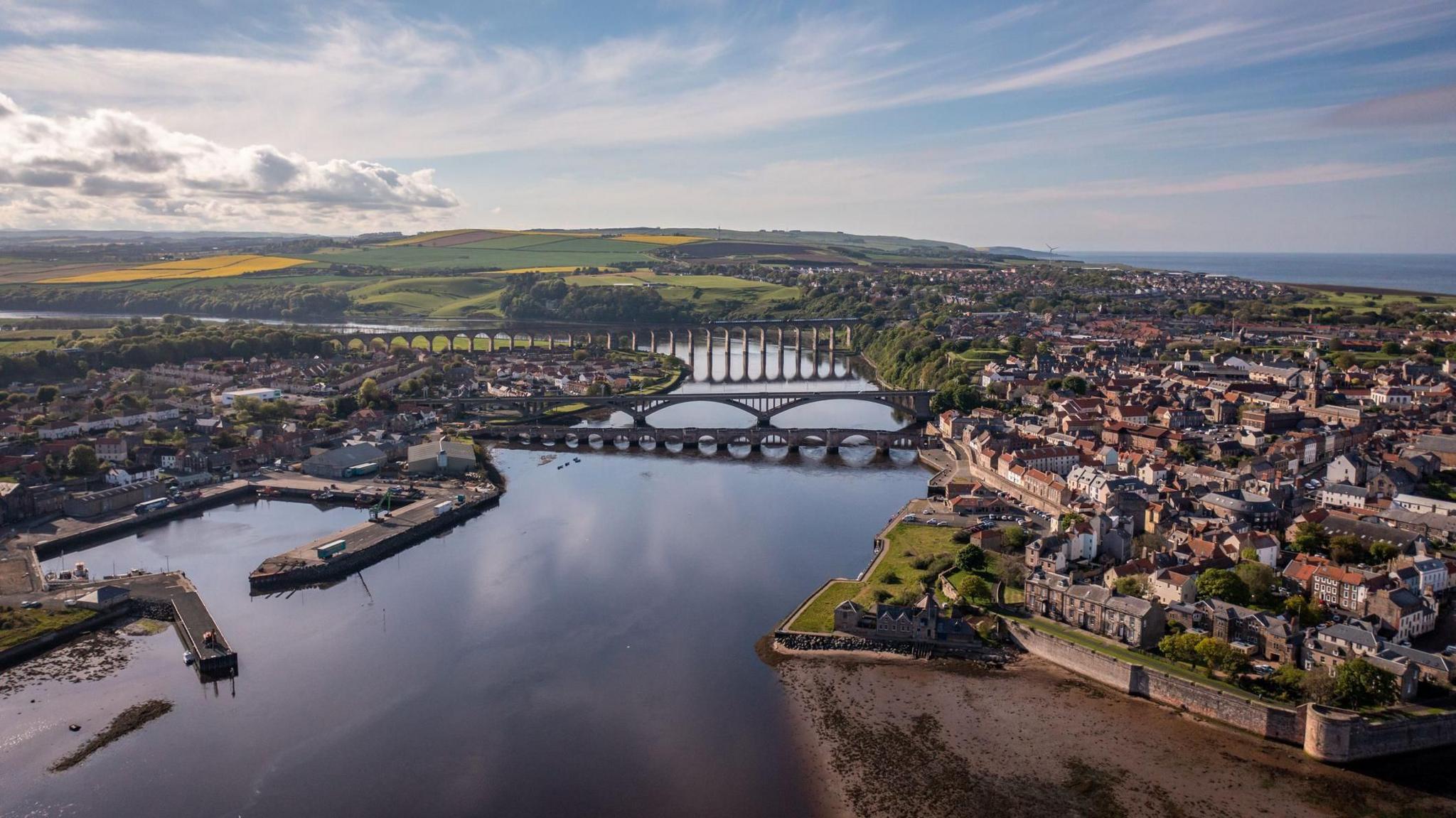 An aerial view of Berwick-upon-Tweed with the river dominating the image with its three bridges over the Tweed. The town stretches on either side with the port to the left. It's a clear blue sunny day.