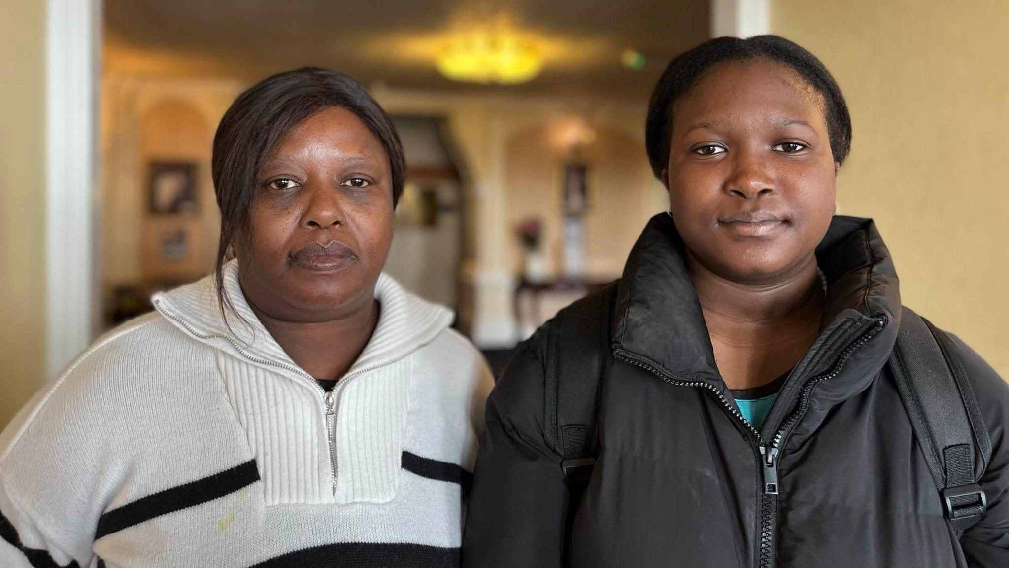 Elizabeth (left, with a white jumper with black stripes on) and Nokutenda (right with a black coat and rucksack) look into the camera