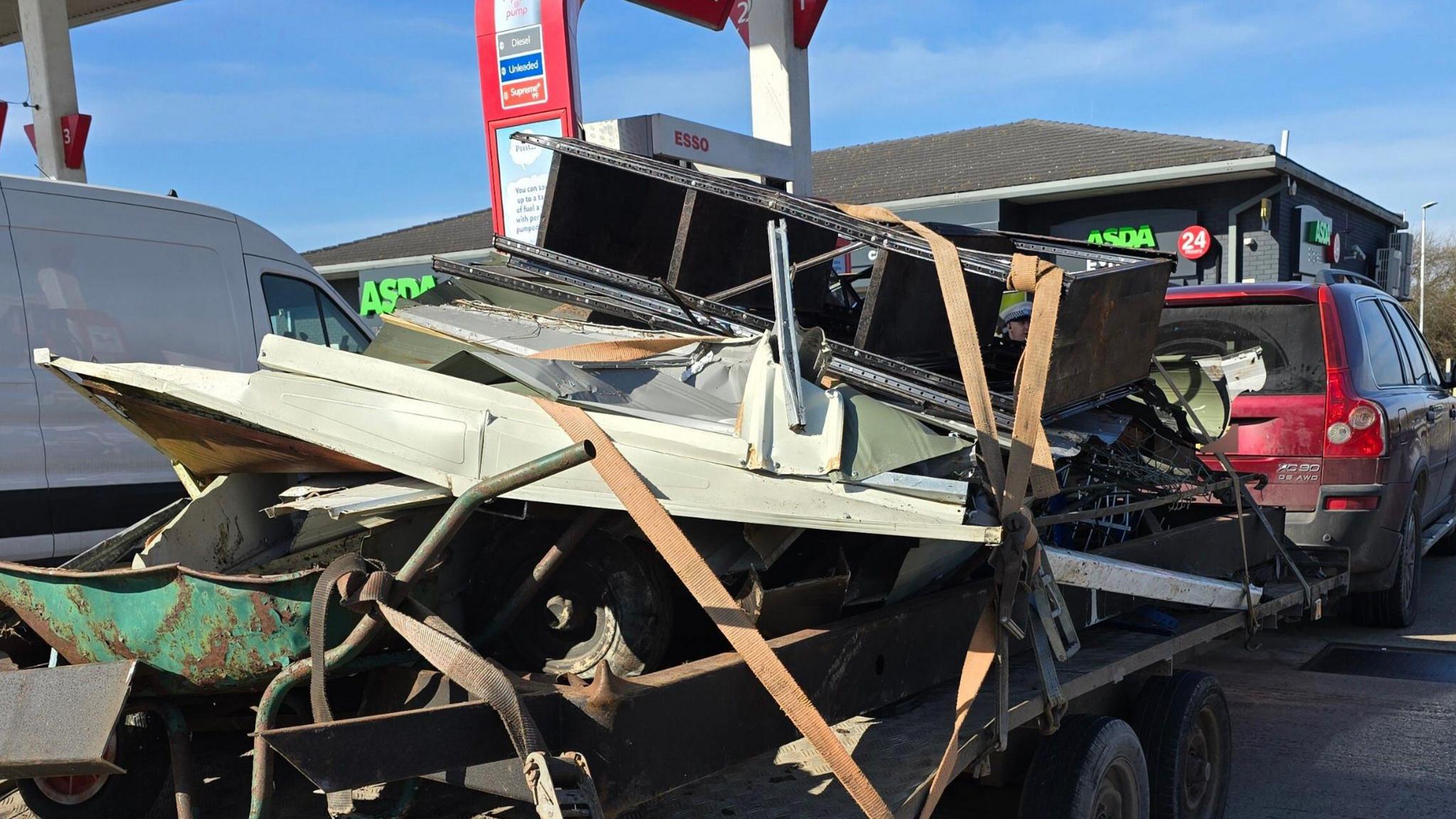 A red car in an Asda petrol station, towing a trailer with various items on it, including what looks like a wheelbarrow and broken down furniture.