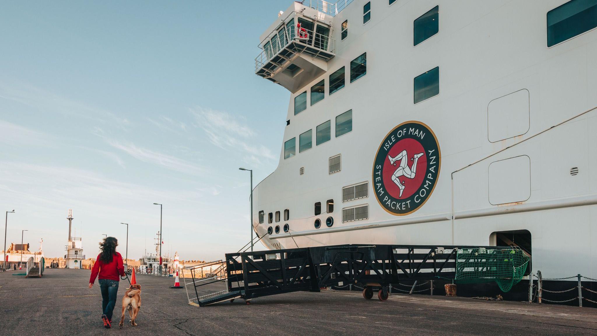 A woman in a red jumper and blue jeans walks a dog on a lead towards a gangaway to get onto a large white ferry with a black and red Isle of Man Steam Packet Company logo.