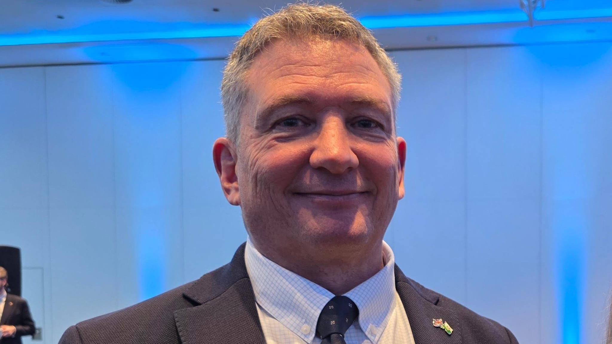 Derek Roberts is wearing a dark suit and a white shirt.