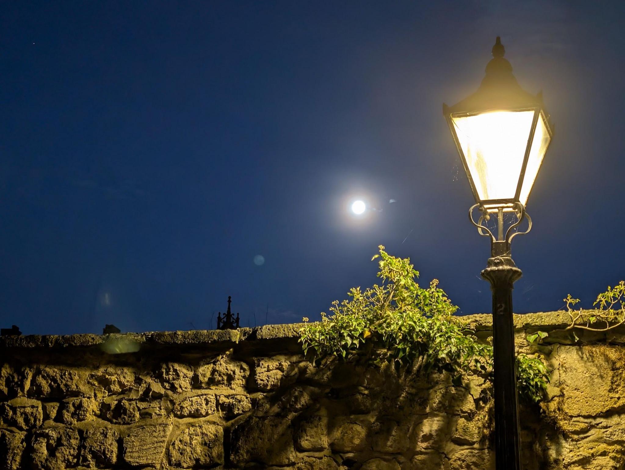A stone wall with an old-fashioned street light against the night sky. The moon is visible and glowing brightly.