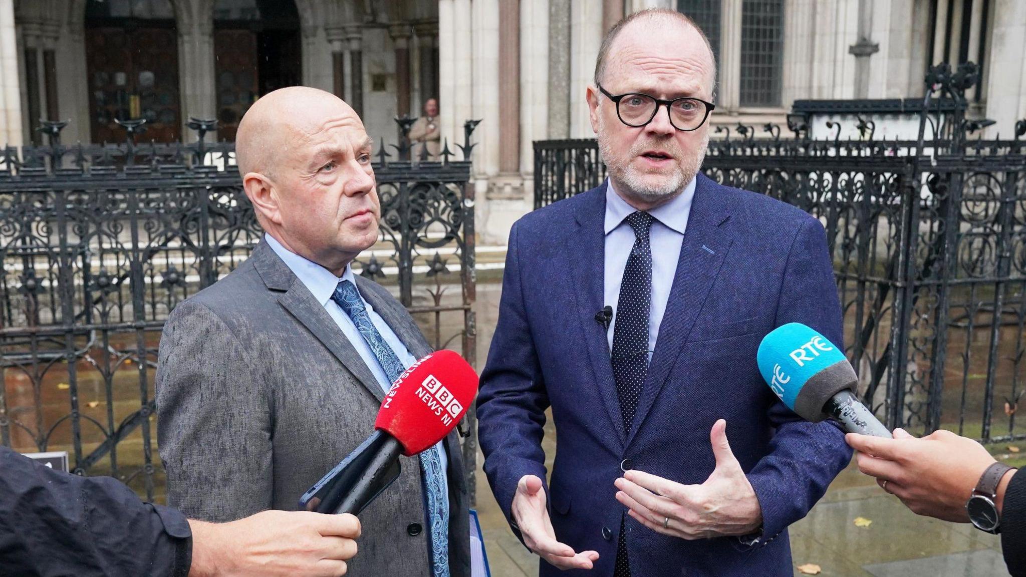 Barry on the left is bald, wearing a grey suit, blue shirt and blue tie.

Trevor on the right is taller, has a bit more hair than Barry and is wearing black round glasses. He is wearing a blue shirt and dark blue navy tie under a dark navy suit. 
 They are being interviewed by news media and microphones are visible, behind them is the Royal Courts of Justice