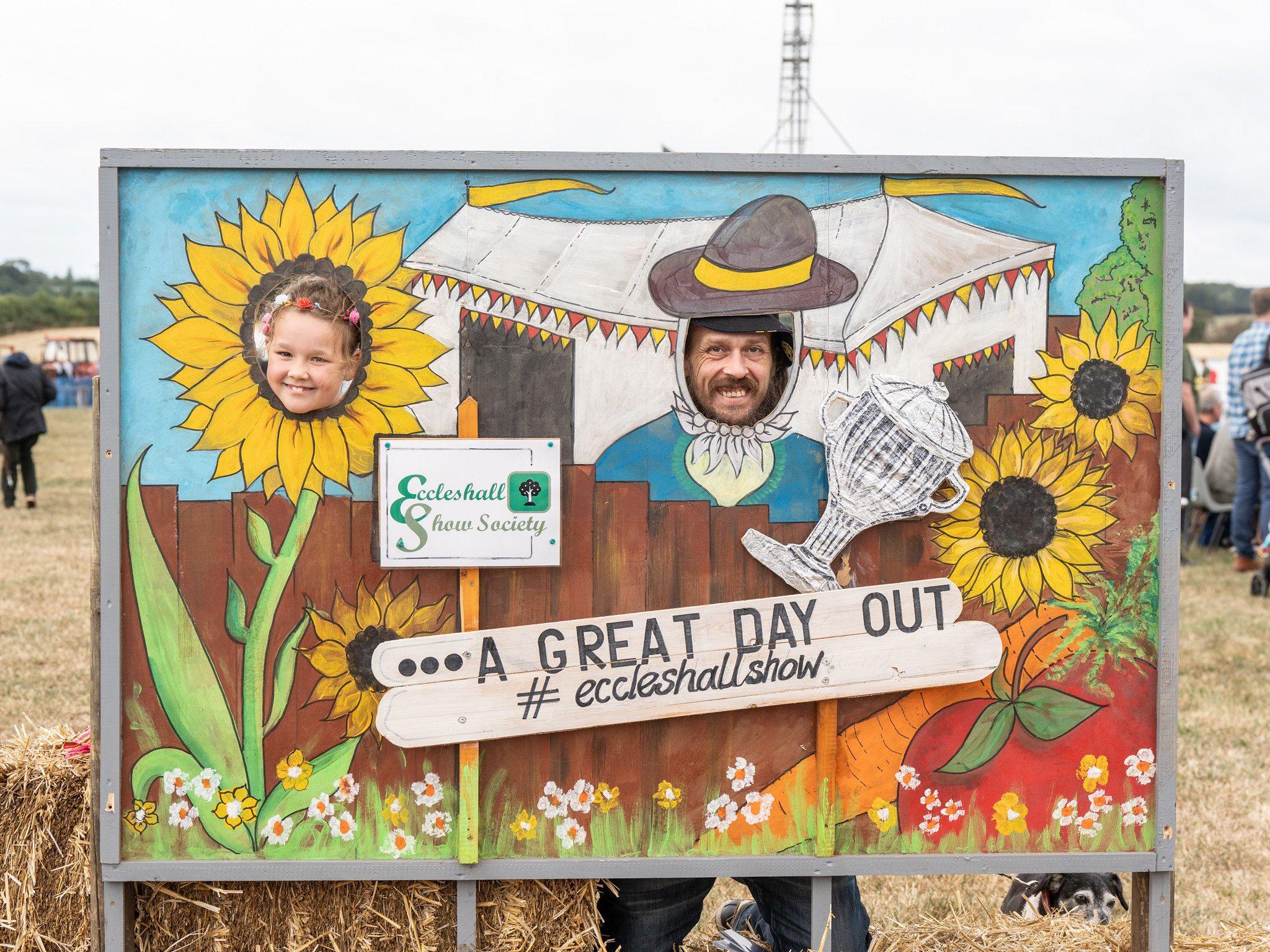 A girl and a man are photographed with their heads peaking through a painted scene showing sunflowers with a sign reading A Great Day Out 