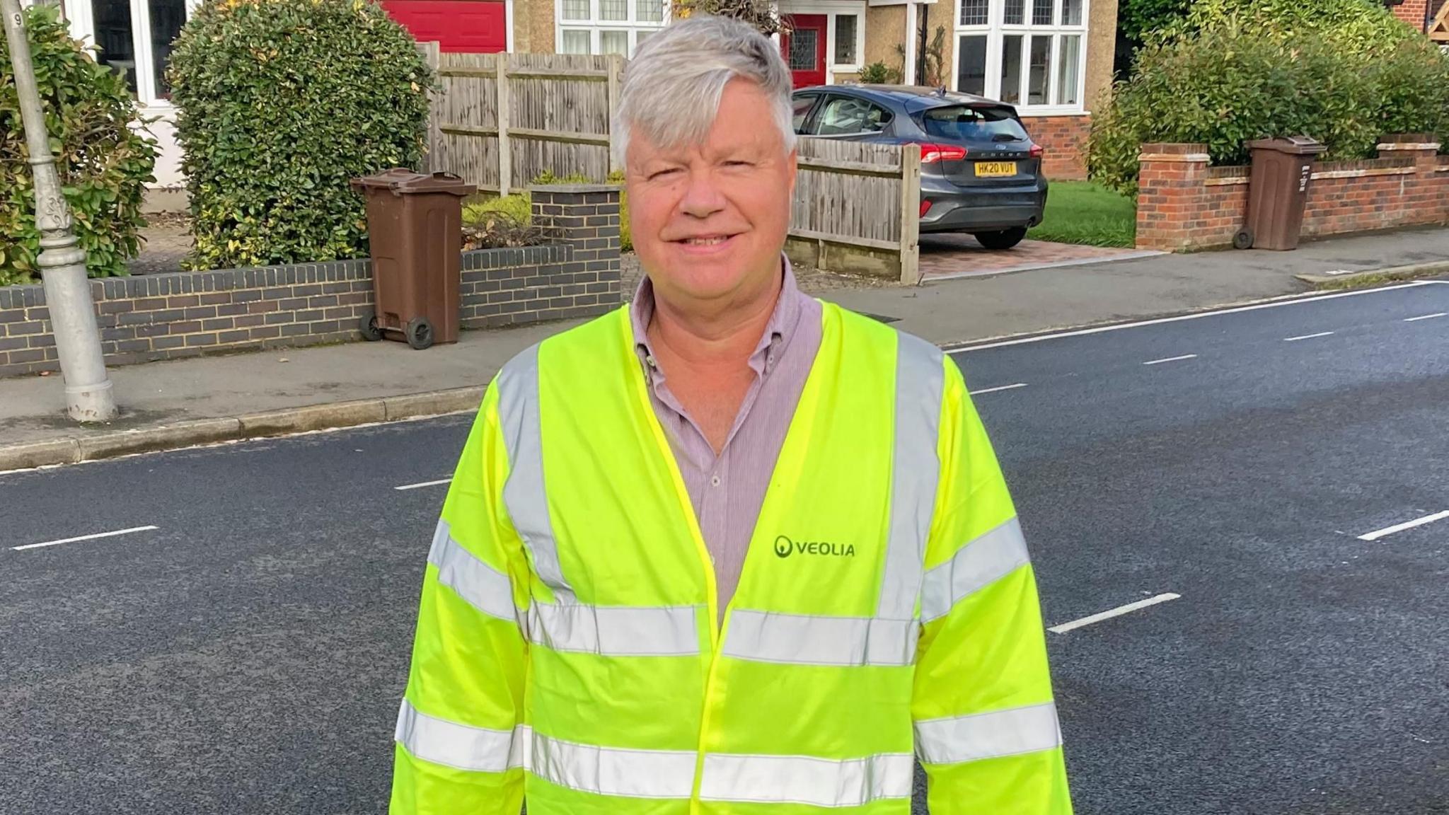 Mark Southgate, wearing a green hi vis jacket and a striped shirt, stands in a residential street. He has grey hair and is smiling.