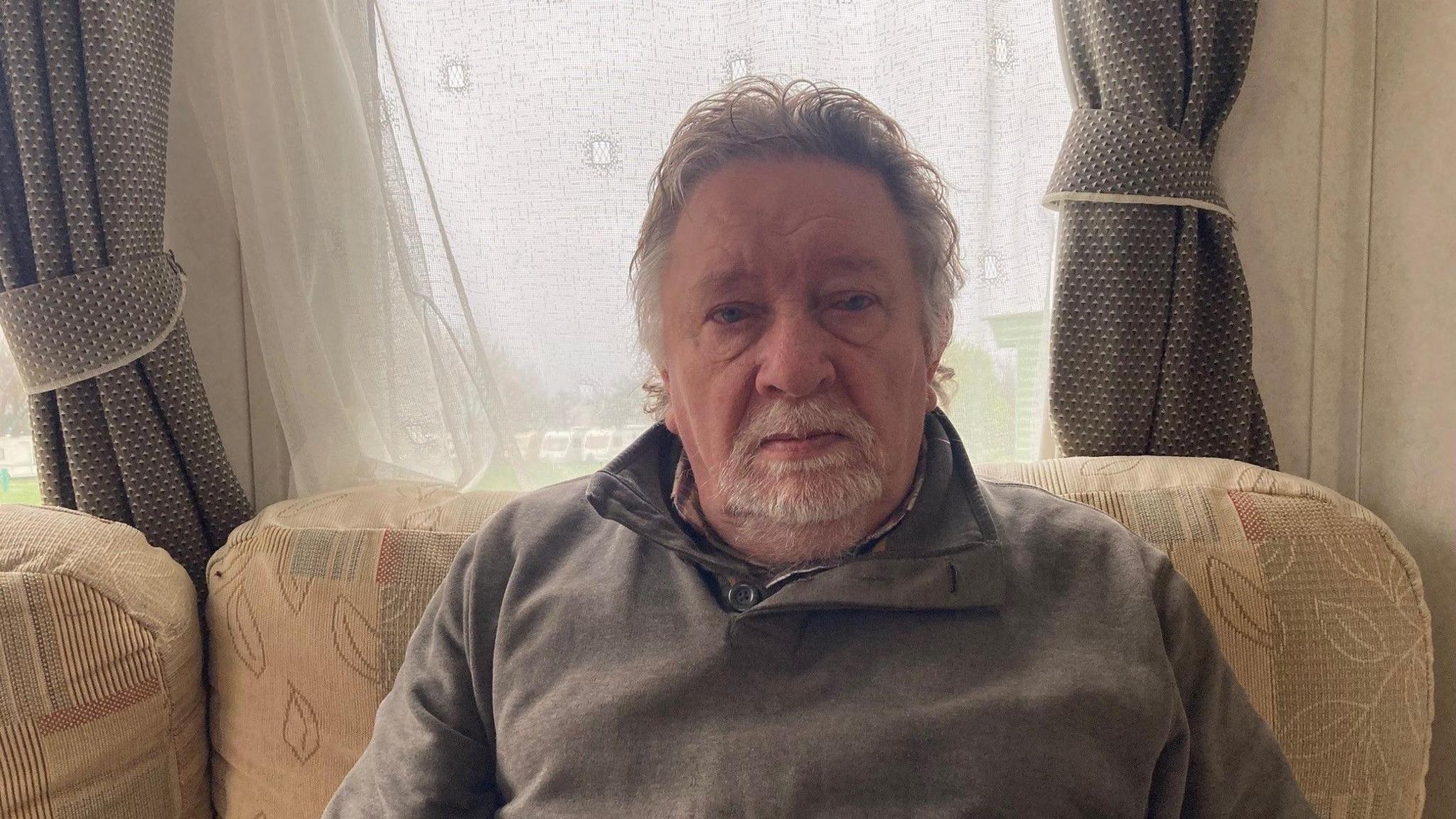 A silver-haired man wearing a grey/brown top sitting inside a caravan. He is sat on a cream patterned sofa in front of window. There is a net curtain across the window and grey patterned curtains pulled back.