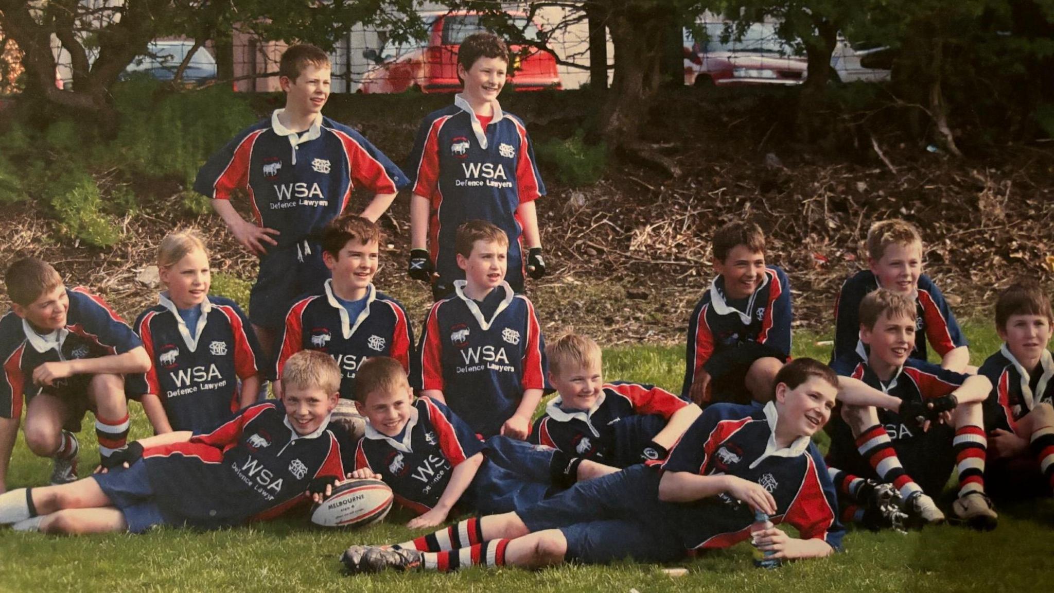 A mini-rugby team (8/9 year olds) in mainly navy kit, with red flashes down side of tops and white, blue and red striped socks, sit and lie on the ground after a game, with blond-haired Sarah Robertson, pictured second left, the only girl amongst the boys.