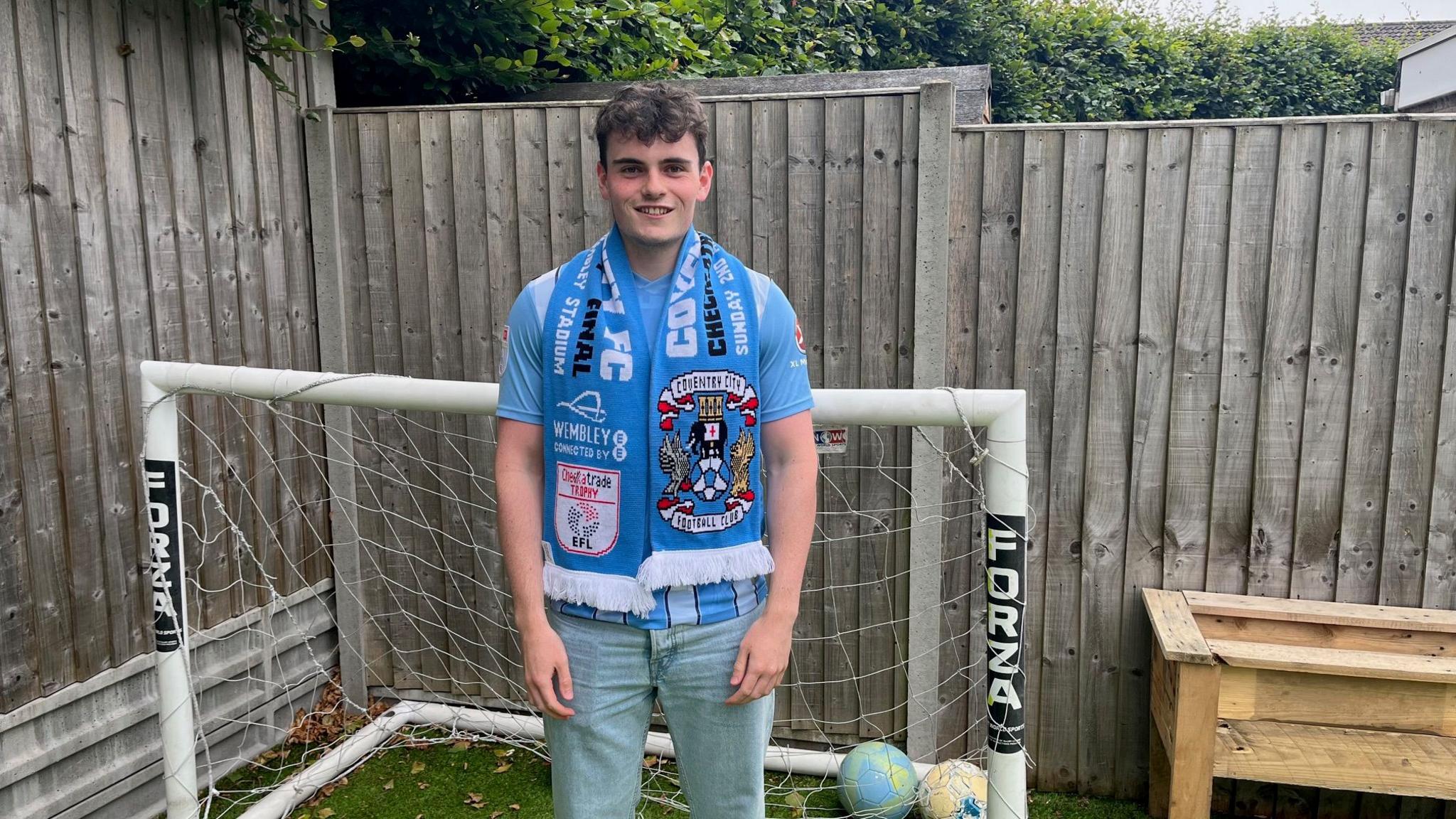 A man stands in in his garden. He is wearing a Coventry City FC t-shirt and jeans as well as a scarf. Goal posts and footballs can be seen on the ground behind him.
