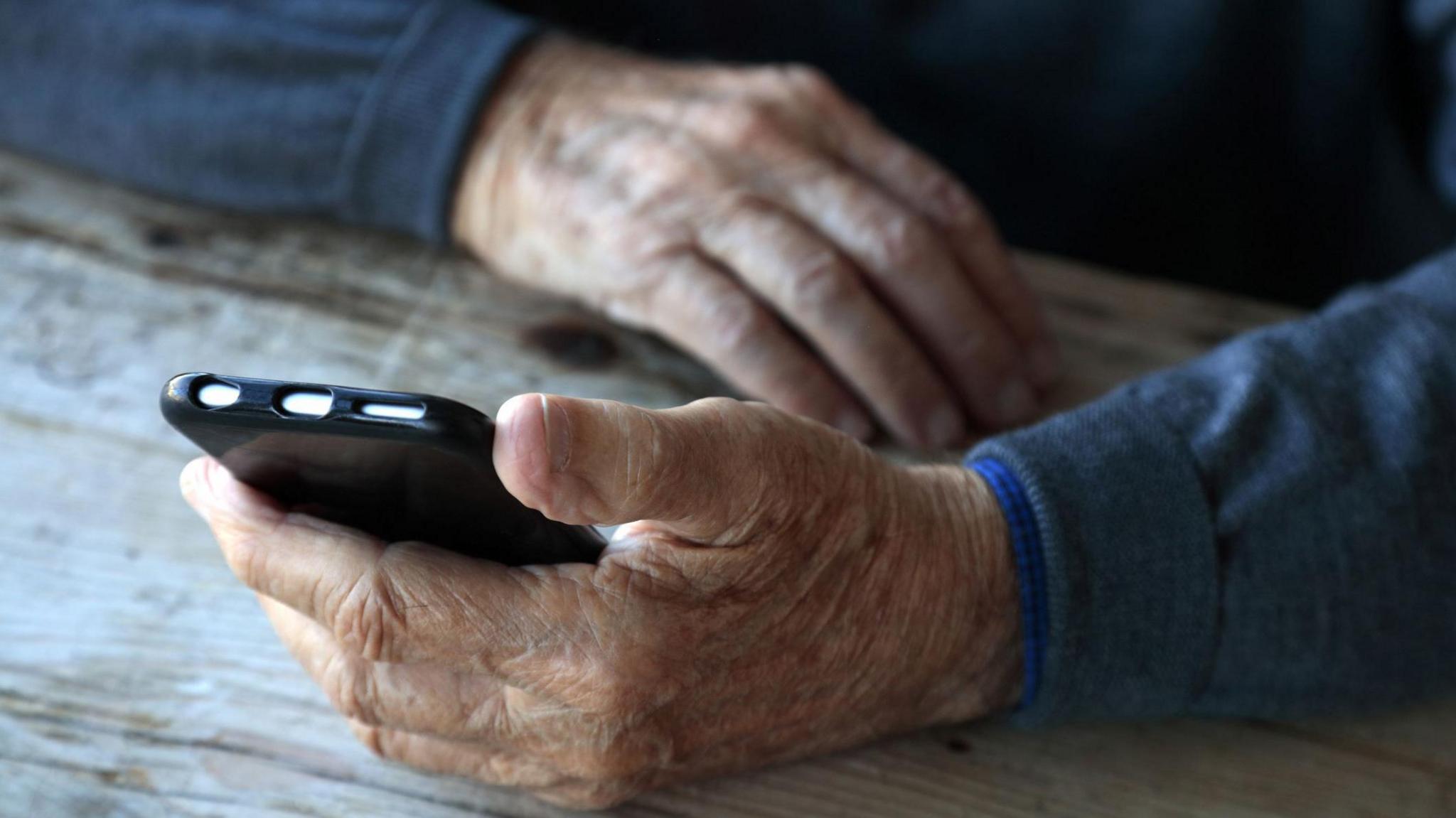 An older man holds a mobile phone in one hand while his other hand rests on a wooden table. He is wearing a blue jersey.