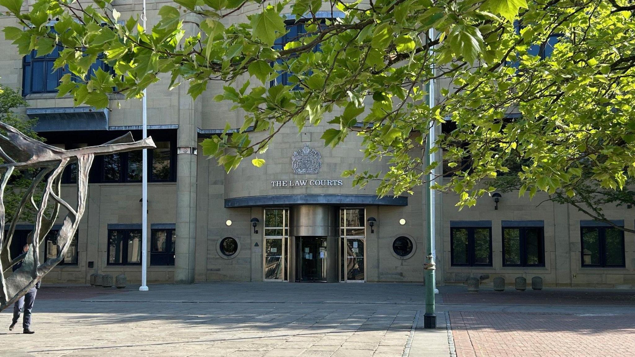 Stone-fronted facade of Bradford Crown Court framed by the branches of a tree in full leaf, with part of a sculpture visible