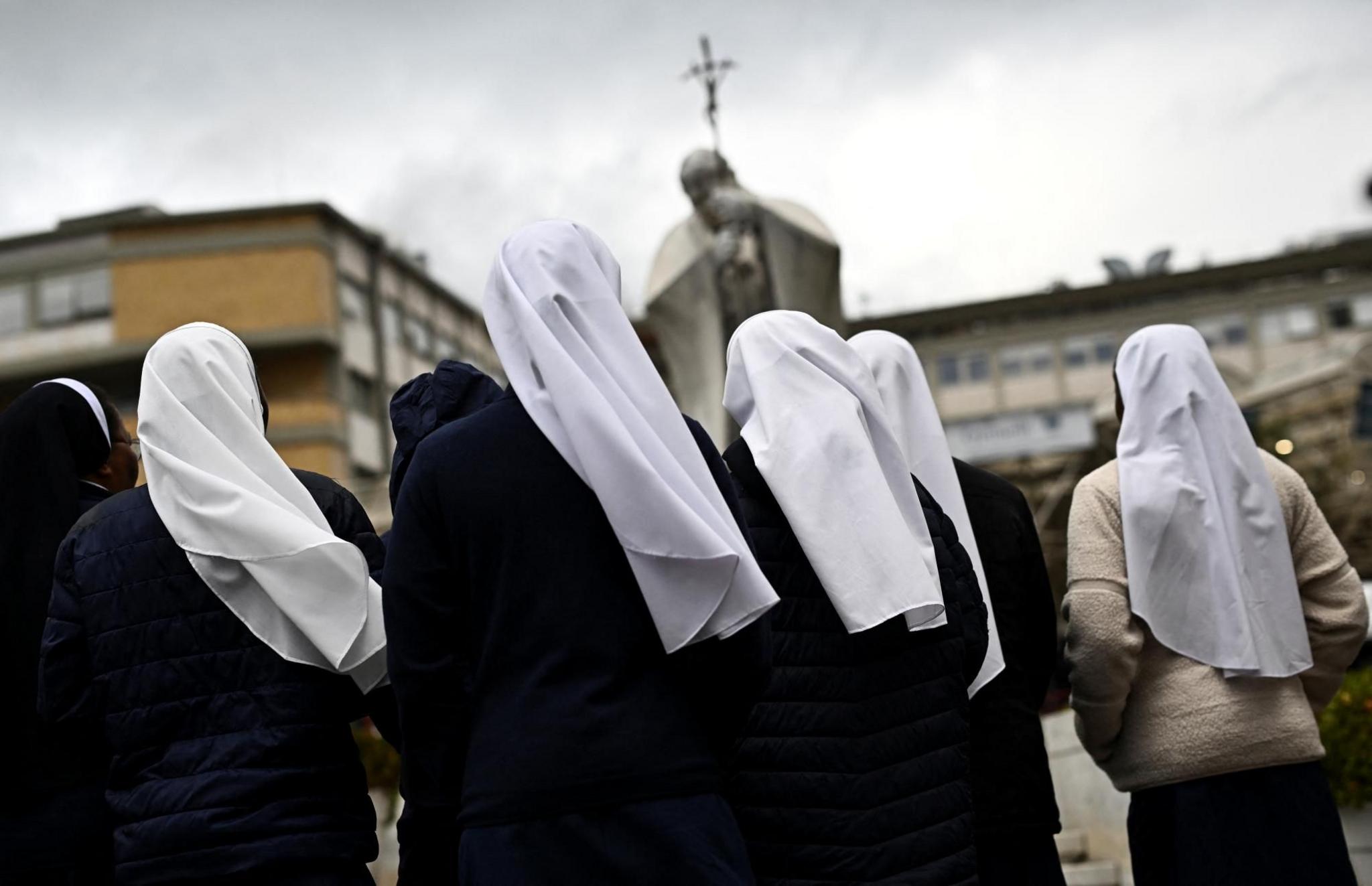 A group of nuns wearing white veils are seen from behind as they pray in front of a statue of Pope John Paul II 
