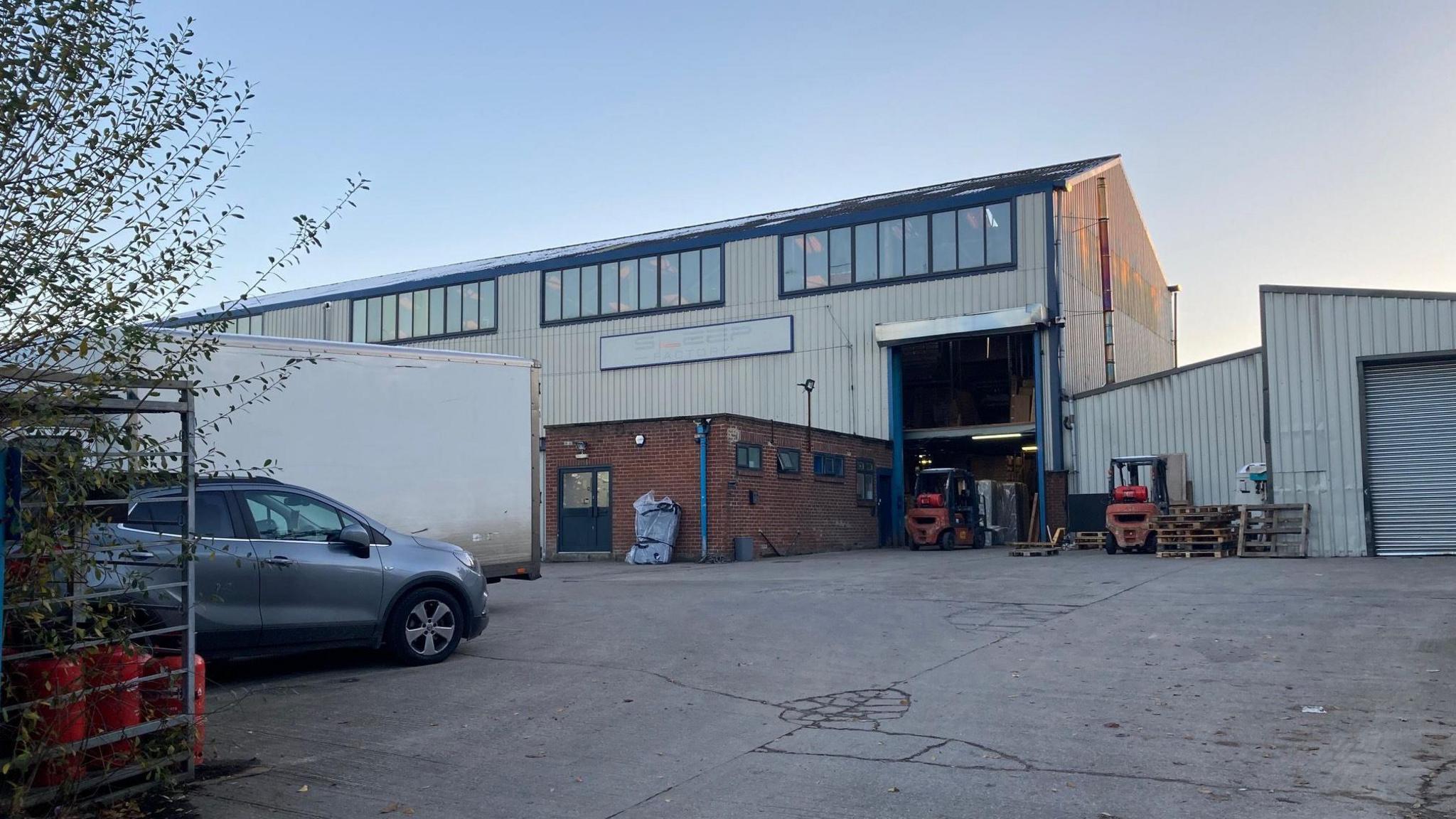 An industrial warehouse building with a large outside yard. A grey car is to the left, while small forklift trucks and building materials sit outside the premises itself.