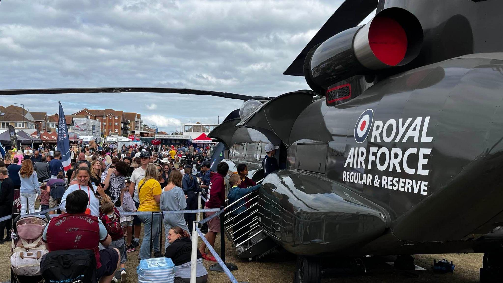 A dark green Chinook helicopter is parked on a field in Clacton. A large crowd of people are gathered outside it, with some queuing to get in. "Royal Air Force regular and reserve" is written in white lettering on the side of the aircraft.