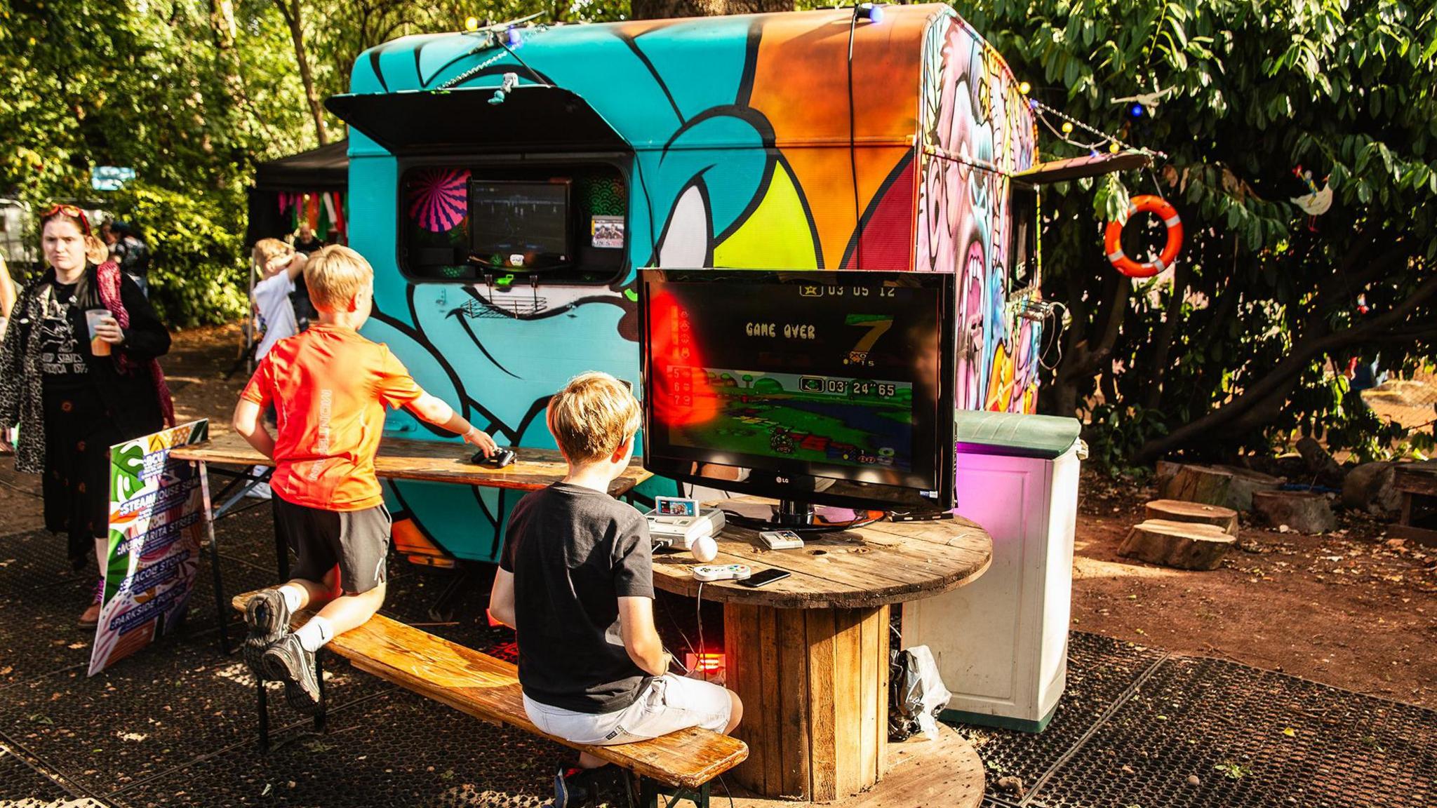 Two young boys on a bench play electronic games on TV screens. 