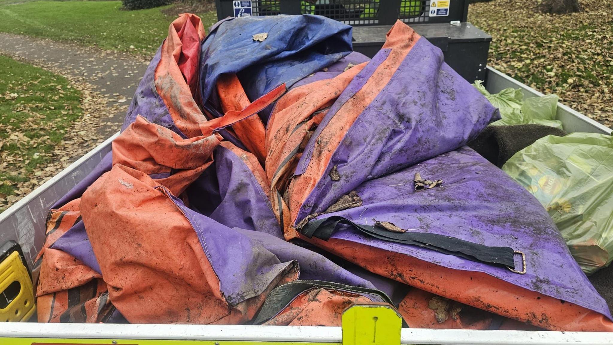 A muddy purple and orange deflated bouncy castle in the back of a truck, next to two bin bags of rubbish