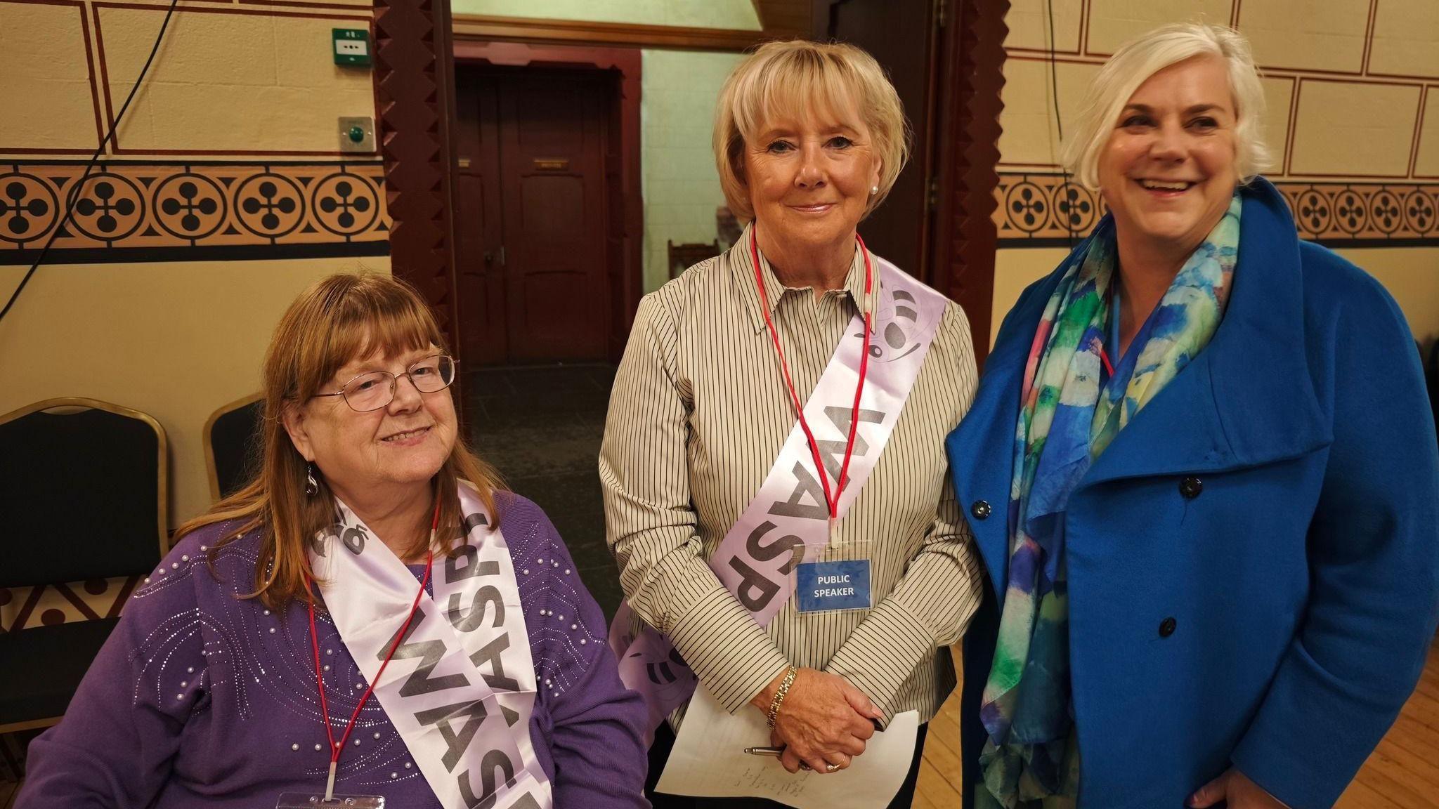 Three women are side-by-side. From the left there is a red-headed woman in a purple jumper. She is next to a blonde woman in a yellow shirt with black trim - they are each wearing a pink sash with "WASPI" on them. To the right is another blonde woman wearing a blue coat with a multi-coloured scarf.