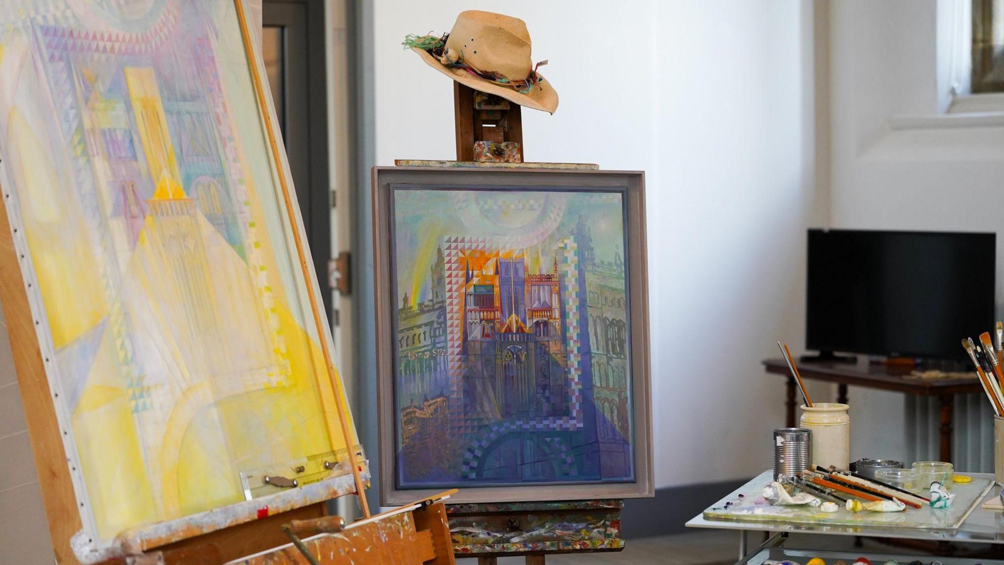 Two easels with two paintings of buildings. On top of the easel on the right is a straw hat. To the right there is a table with various paintbrushes and paint. There is a TV in the background.