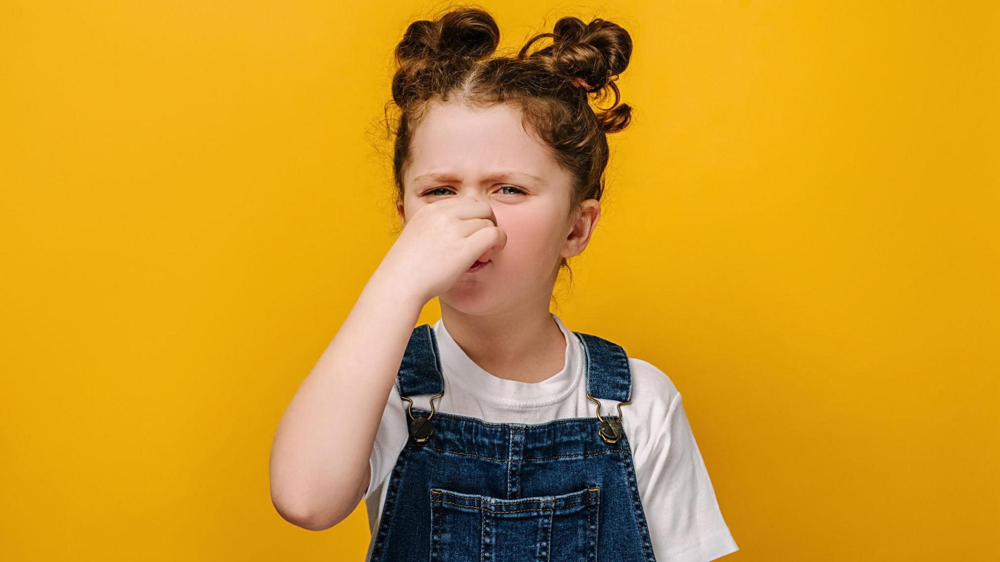 A young girl wearing denim dungarees holds her nose as if she has smelled something unpleasant. 