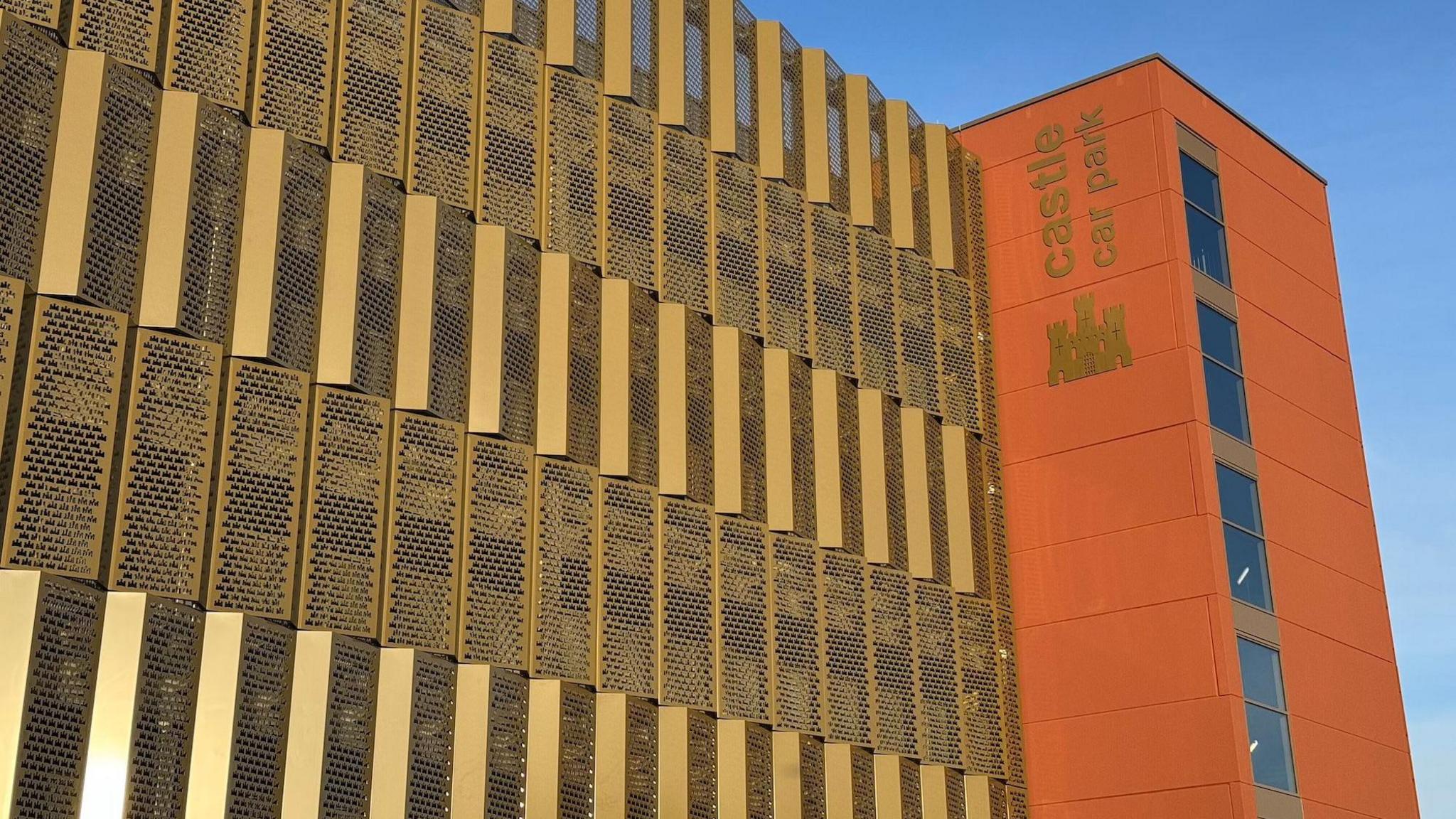 A multi-storey car park, with gold panelling and a terracota-coloured cladded stairwell to the right. There is a gold image of a castle underneath lettering which reads Castle Car Park.