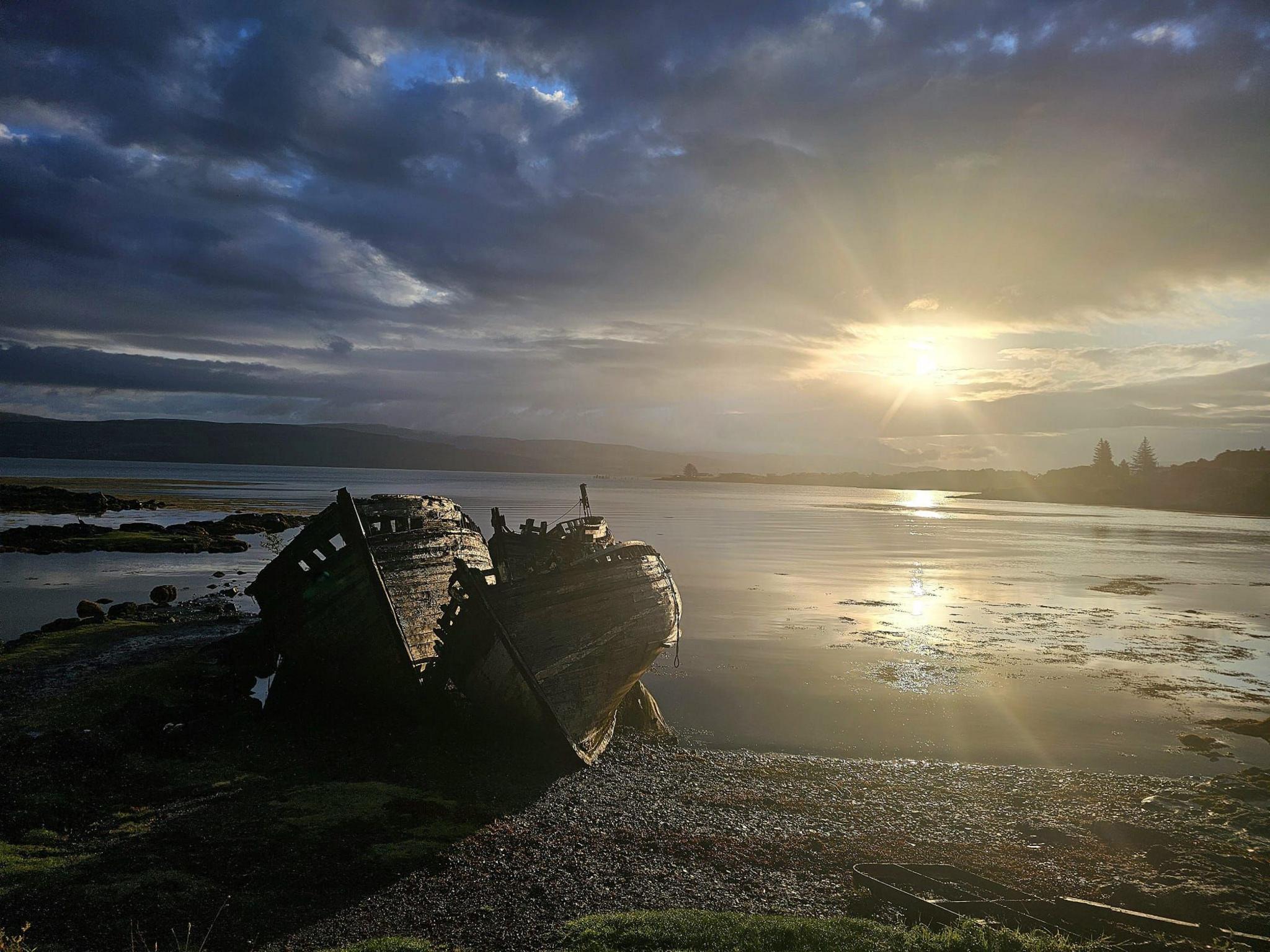 An abandoned boat on the shore next to a loch, with the sun breaking through dark clouds above it