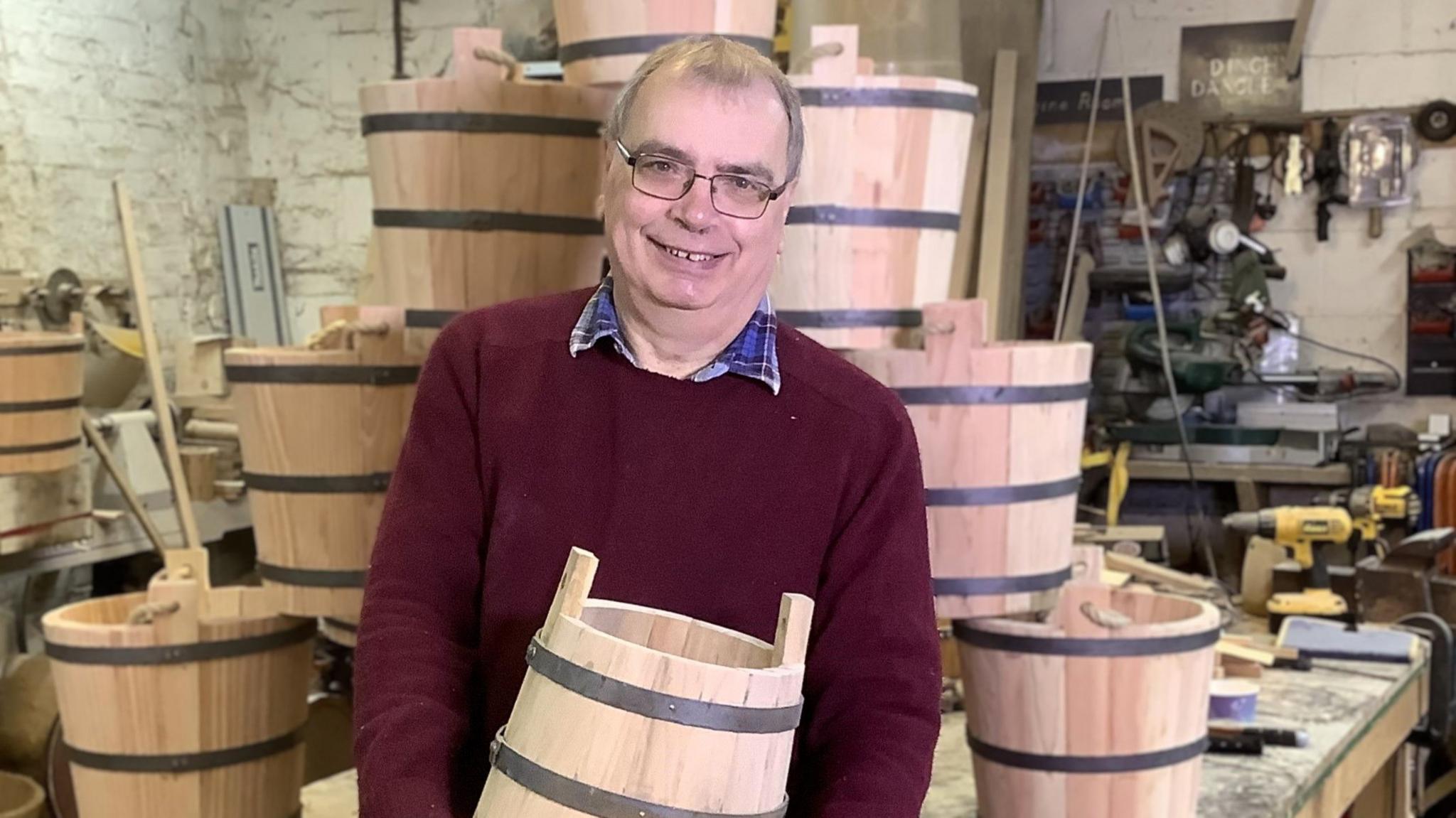 Alan Paulus wearing a red jumper and glasses. He is holding a bucket and is stood in front of a pile of buckets in his workshop.