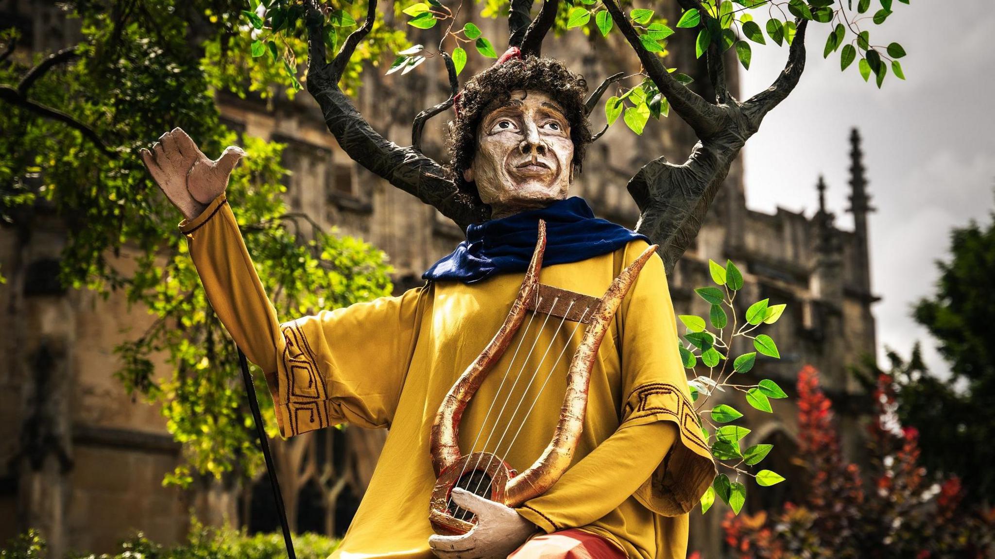 A puppet of Orpheus, taken in front of Gloucester Cathedral. He wears a yellow tunic and a blue scarf, and has brown curly hair. He holds a golden lyre in his lap with one hand, while the other is raised.