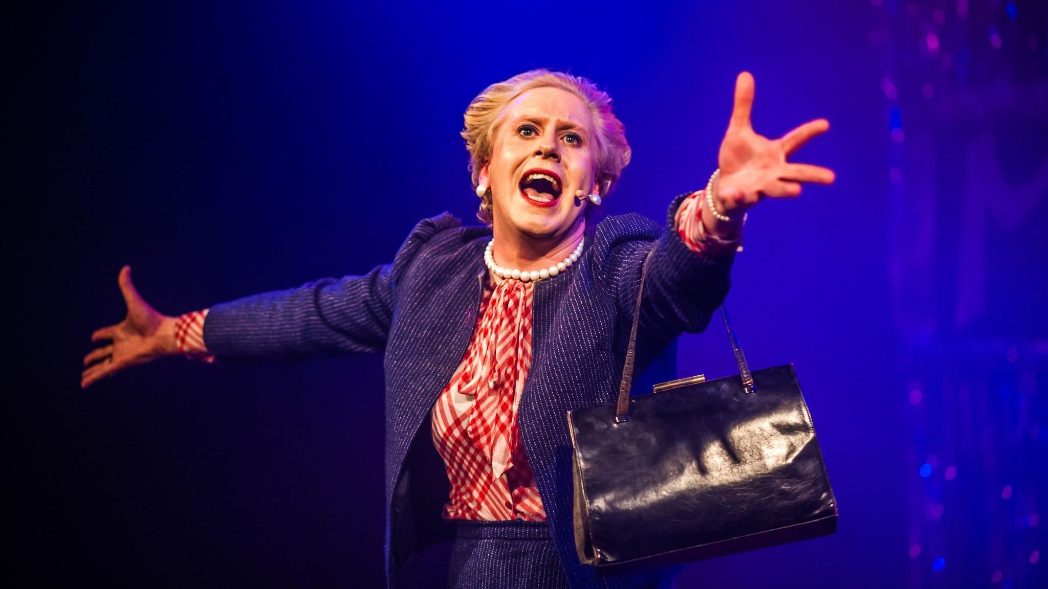 A performer dressed as Margaret Thatcher sings into a small microphone attached to their headset as they fling their arms wide during a performance at Bristol Old Vic. They are wearing a dark blue skirt and matching jacket, a red and white shirt and holding a black leather handbag from their left arm