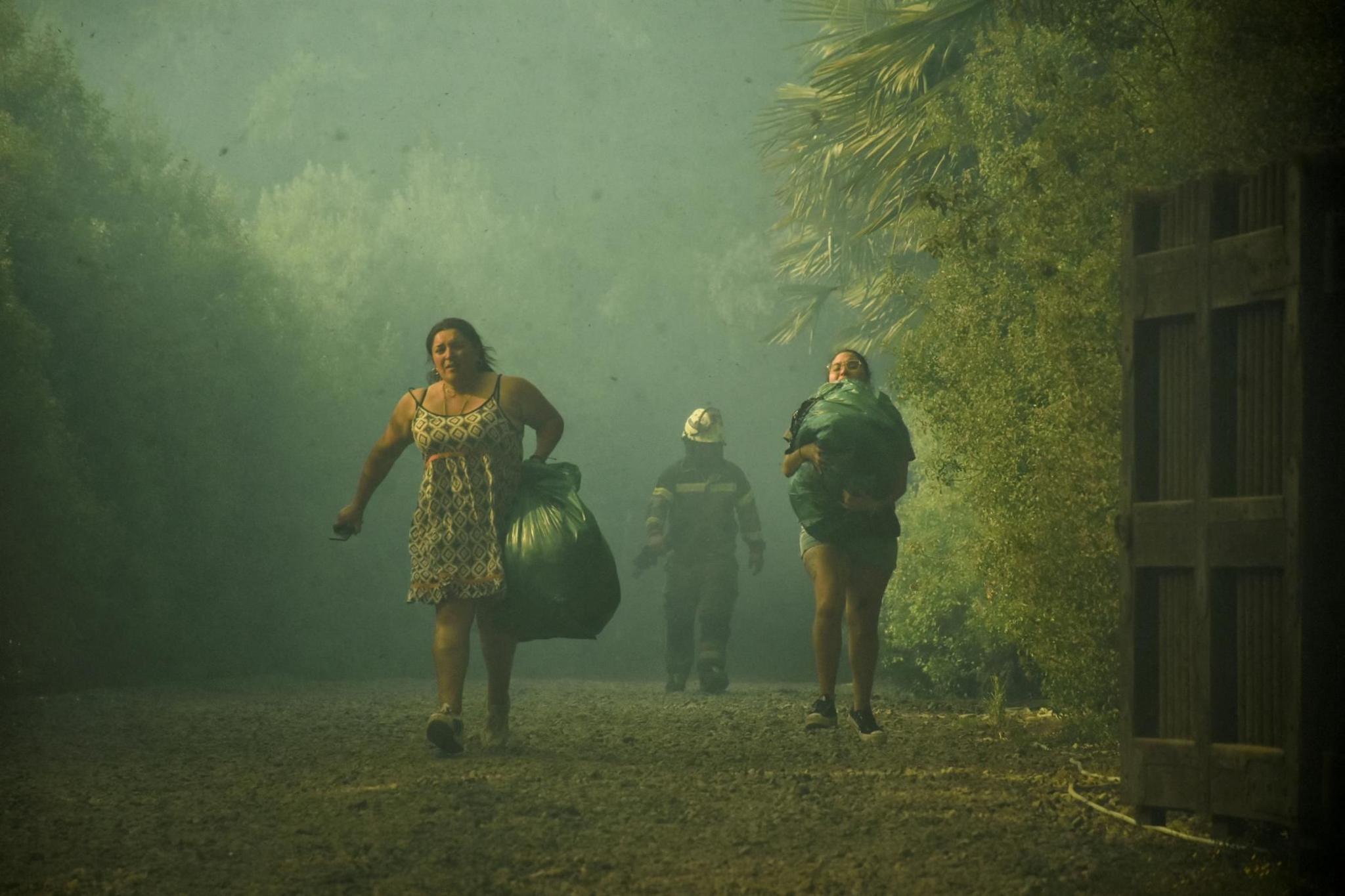 Two women carry their belongings in plastic bags as smoke from wildfire hangs in the air 
