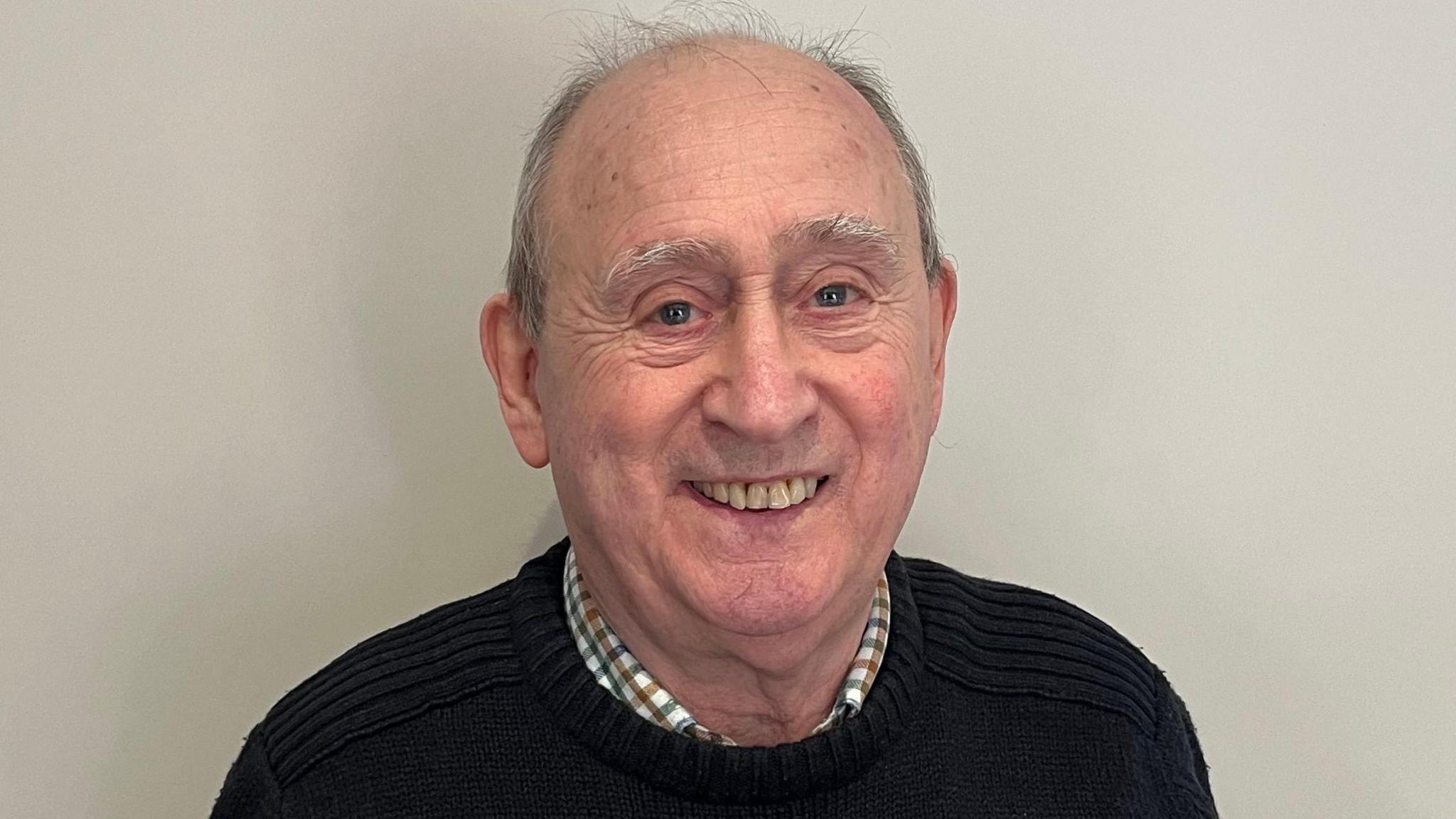 Bill Smith is in the middle of the image looking towards the camera smiling. He has short grey hair and is wearing a black jumper with a shirt underneath.