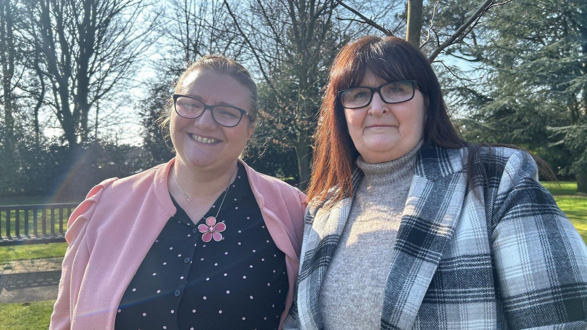 Two women, both with dark hair and glasses, stand side by side in the sunshine. One is wearing a pink cardigan with a flower necklace and a black top with light spots. The other is wearing black-and-white shirt on top of a beige jumper.    